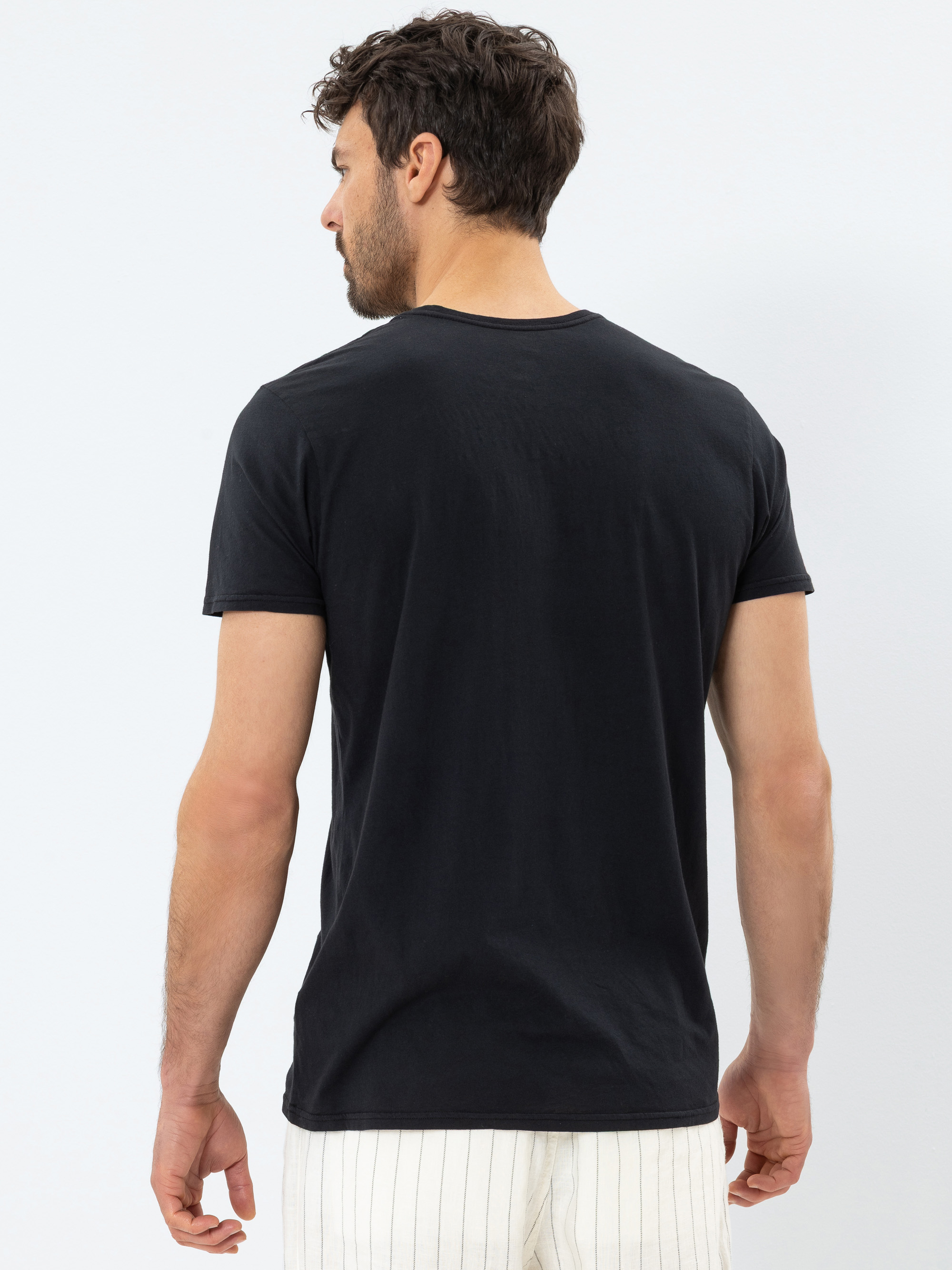 Ein Mann mit kurzen, dunklen Haaren steht mit dem Rücken zur Kamera, trägt ein schlichtes schwarzes T-Shirt und eine weiße Hose mit dünnen schwarzen Längsstreifen vor einem hellen Hintergrund.