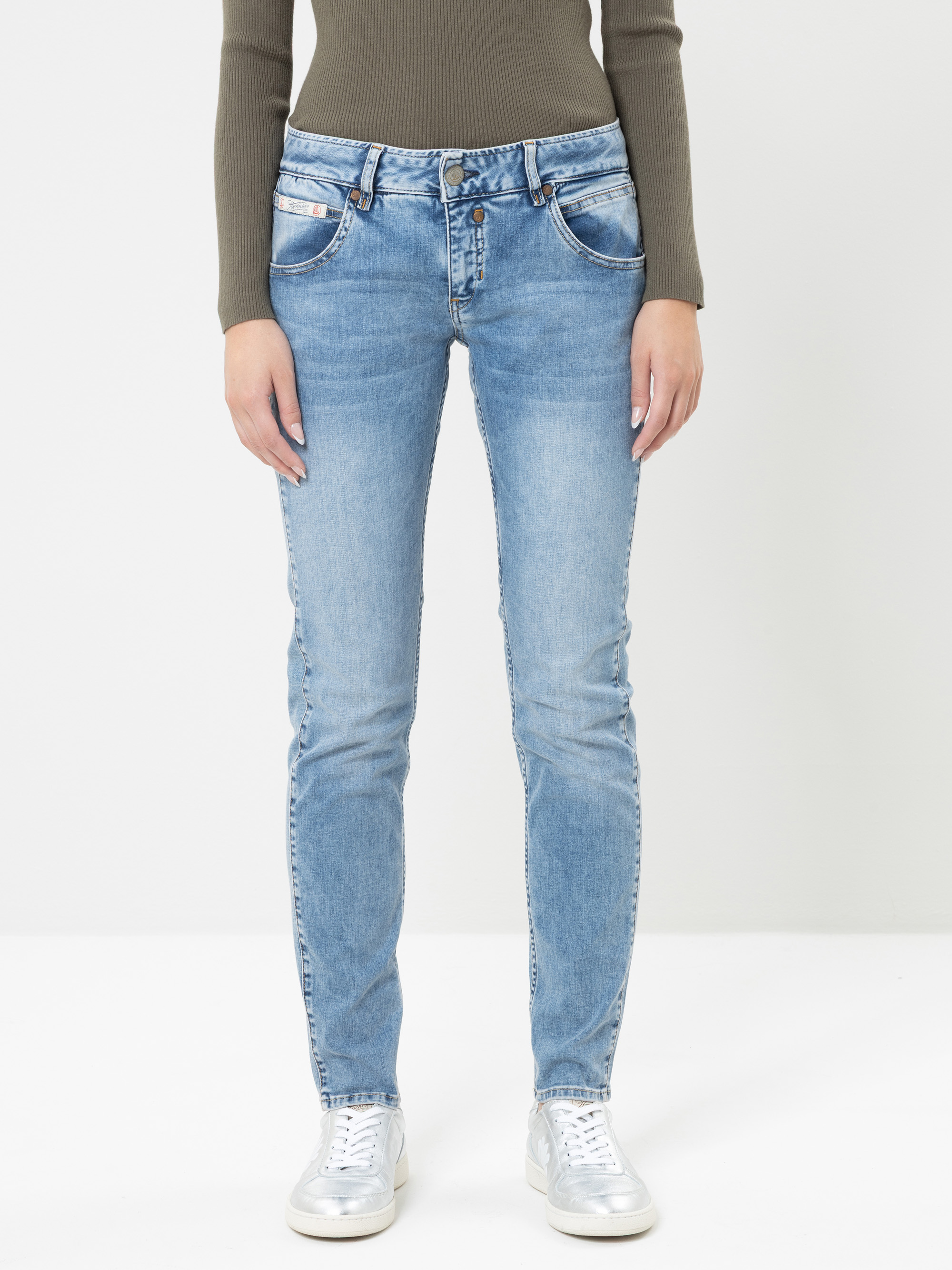 Eine Person, die ein geripptes, langärmeliges, olivfarbenes Oberteil, hellblaue Skinny-Jeans und silberne Sneaker trägt, steht vor einem schlichten weißen Hintergrund.