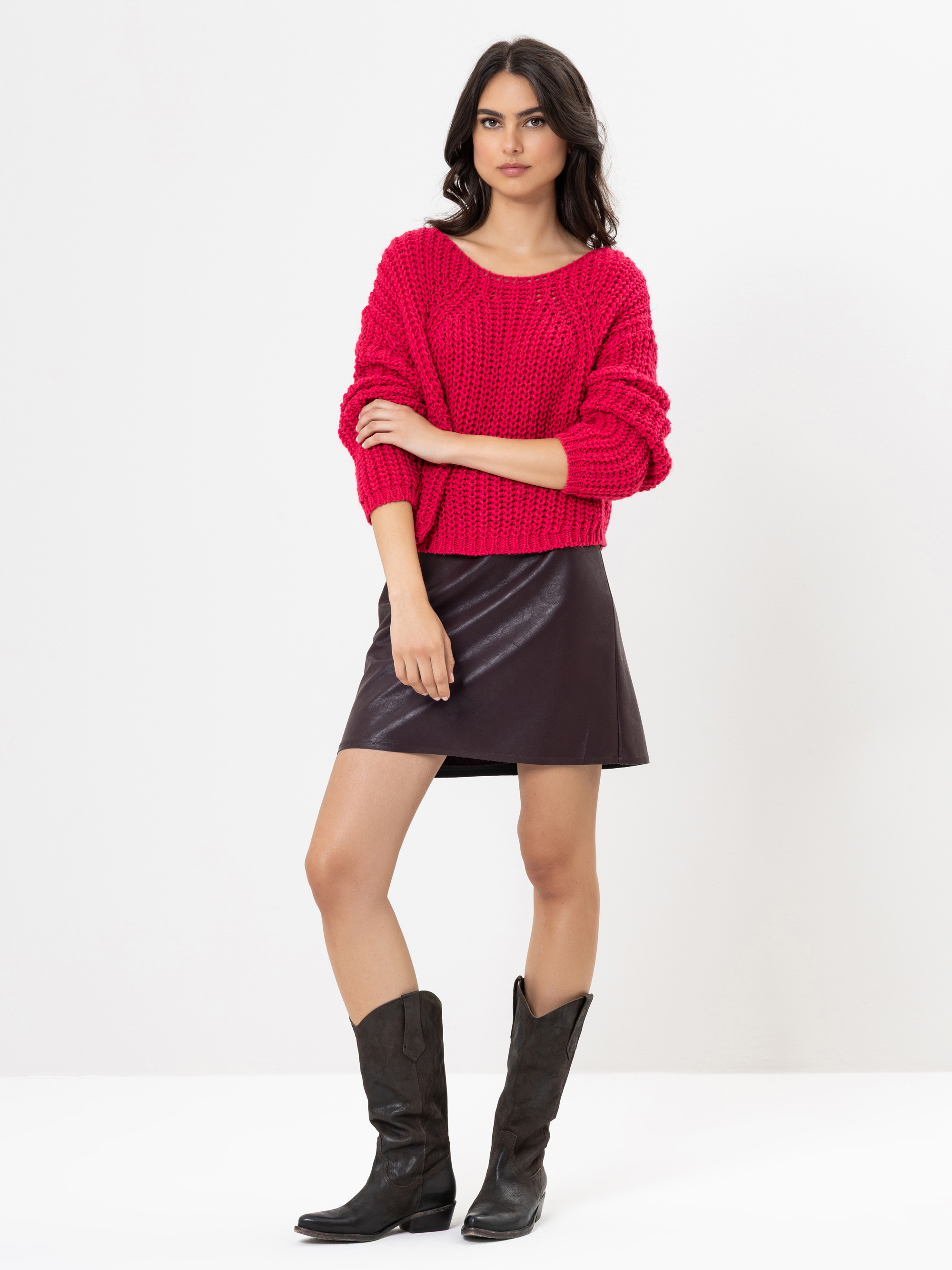 Eine Frau steht vor einem weißen Hintergrund und trägt einen knallroten Strickpullover, einen kurzen dunkelbraunen Lederrock und hohe schwarze Stiefel. Sie hat langes dunkles Haar und eine entspannte, selbstbewusste Pose.