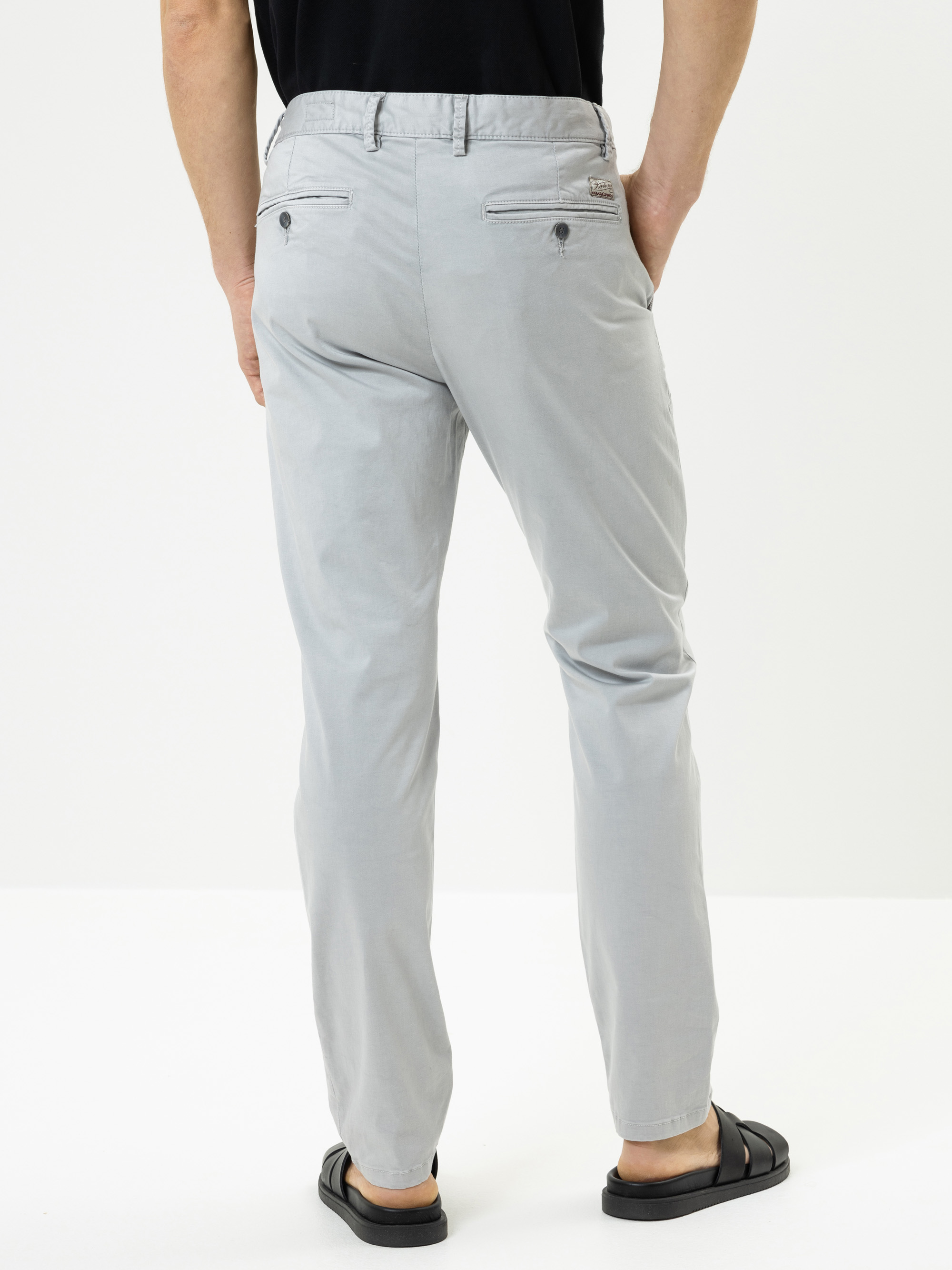 Eine Person, die eine hellgraue Hose, ein schwarzes Hemd und schwarze Sandalen trägt, steht von hinten gesehen vor einem schlichten weißen Hintergrund.