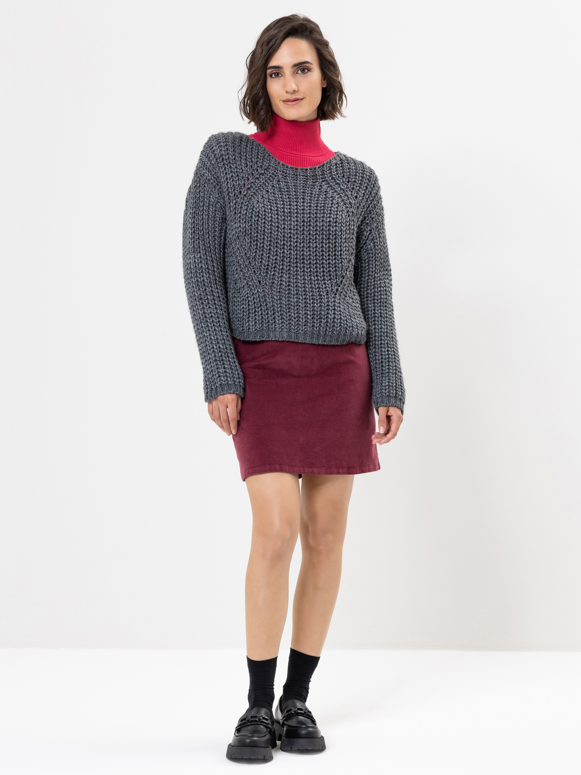 Eine Person steht vor einem weißen Hintergrund, trägt einen grauen Strickpullover über einem roten Rollkragenpullover, einen weinroten Rock, schwarze Socken und schwarze Schuhe und schaut mit neutralem Blick in die Kamera.