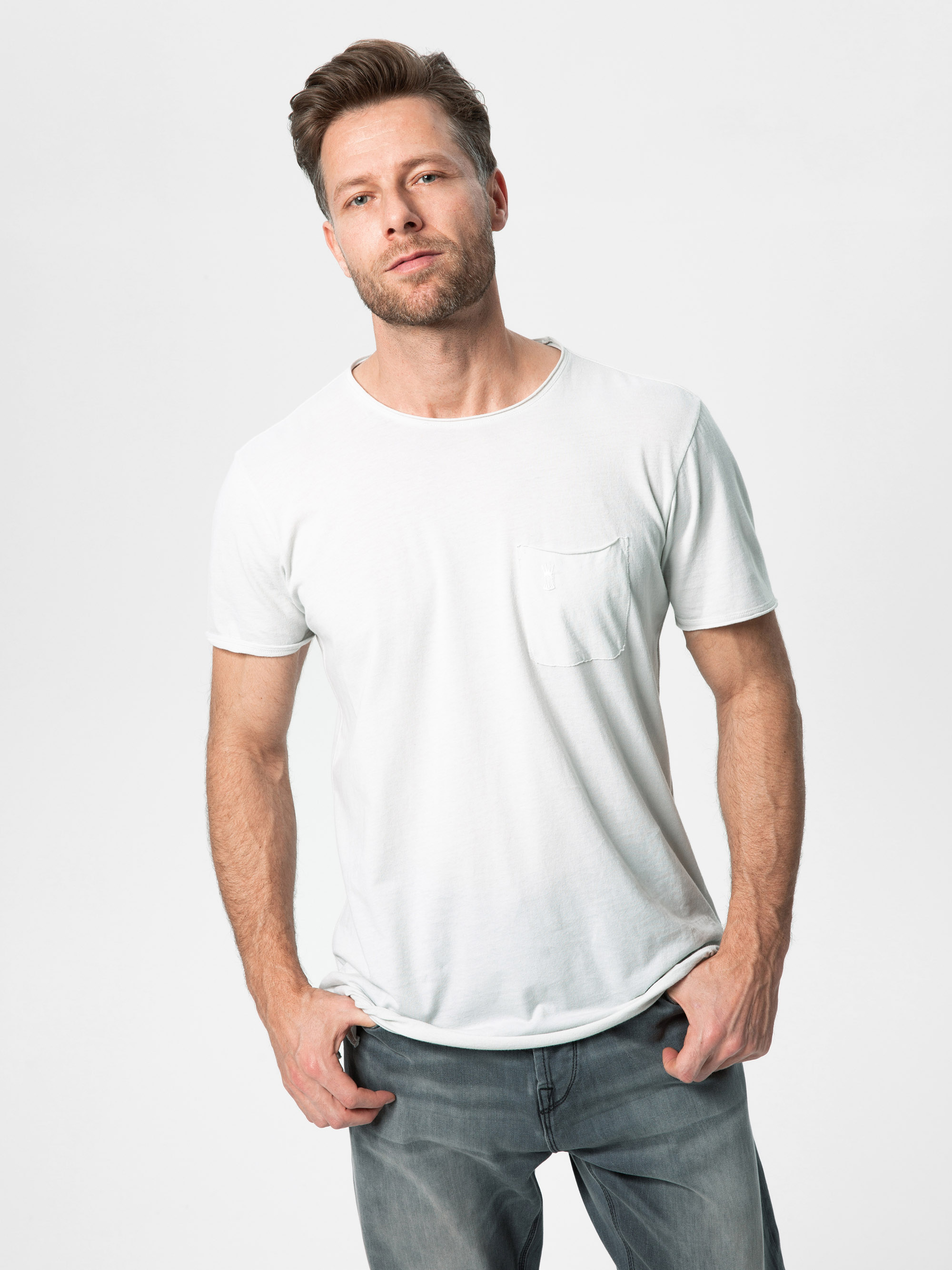 Ein Mann mit hellbraunem Haar und Bart steht vor einem schlichten weißen Hintergrund. Er trägt ein weißes T-Shirt mit einer Tasche und verblichene graue Jeans. Er nimmt eine entspannte Haltung ein, hat die Hände in den Taschen und einen leichten, selb