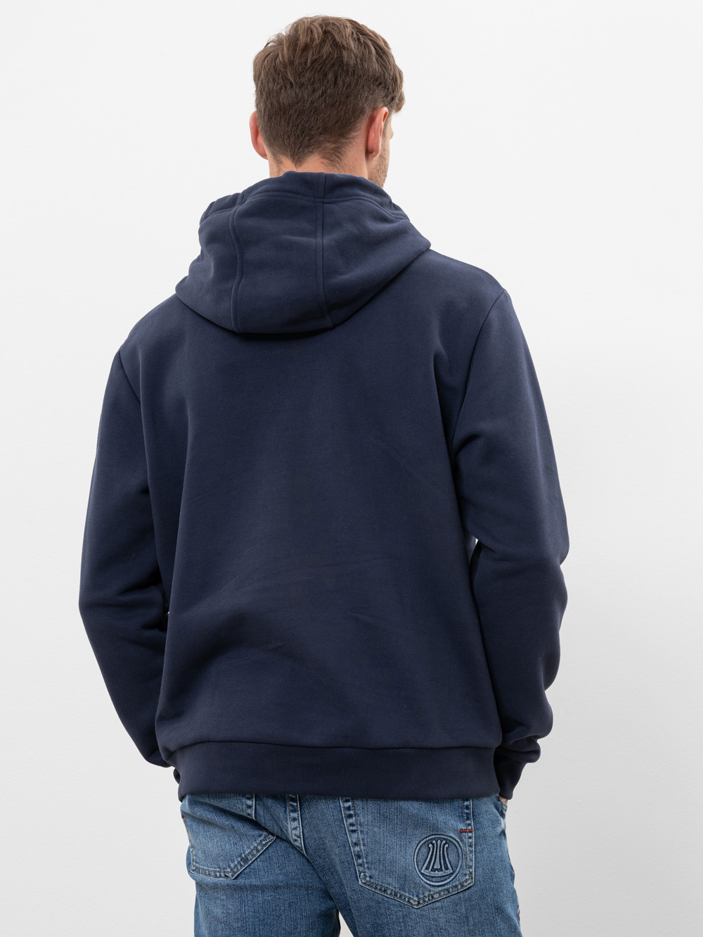 Eine Person, die ein dunkelblaues Sweatshirt mit Kapuze und blaue Jeans trägt, steht mit dem Rücken zur Kamera vor einem schlichten weißen Hintergrund.
