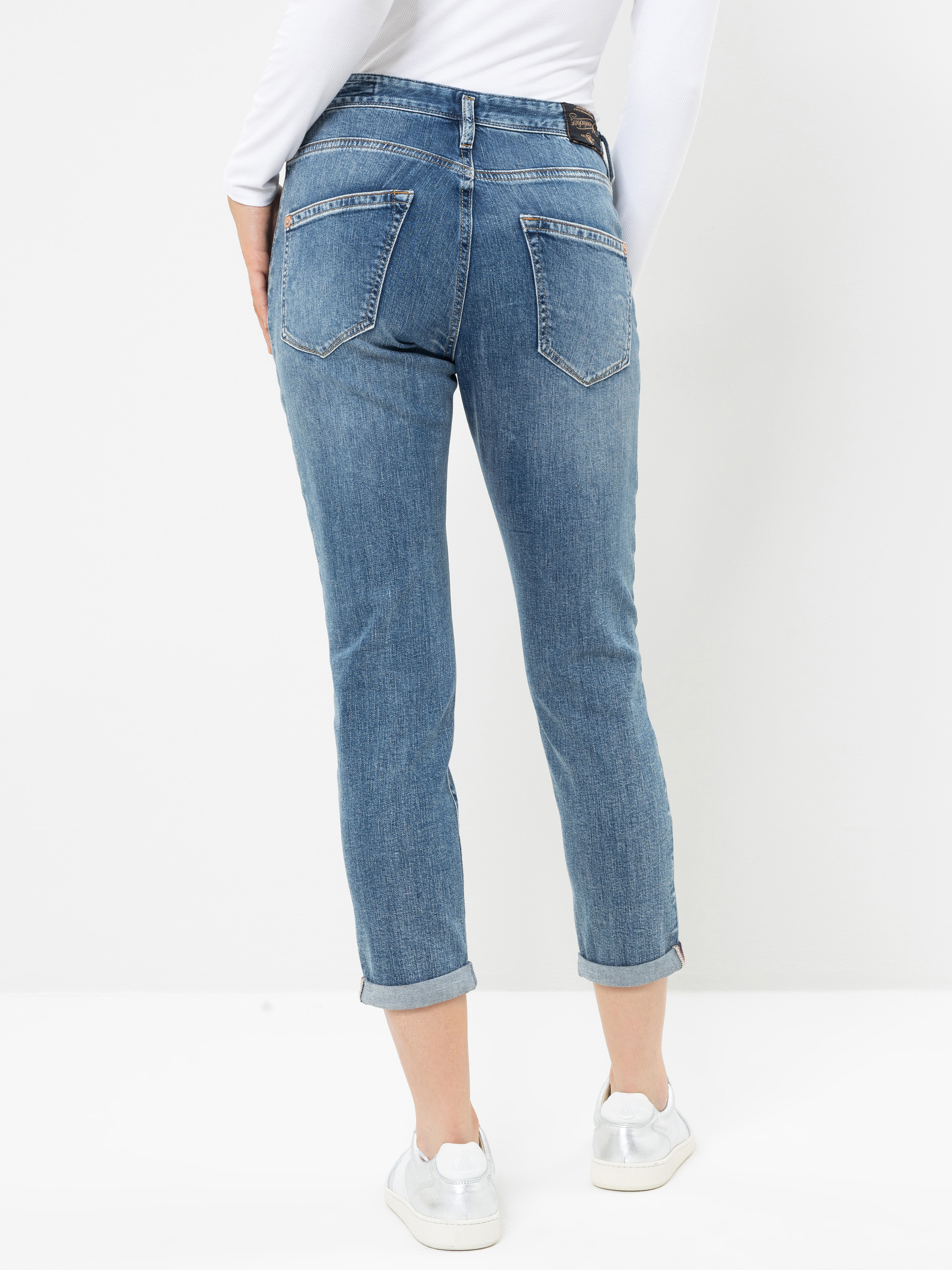 Eine Person in blauen Jeans mit Umschlag und weißen Sneakers steht vor einem weißen Hintergrund. Das Bild zeigt eine Rückenansicht, die die Passform und das Design der Jeans hervorhebt, wobei beide Hände an den Seiten ruhen.