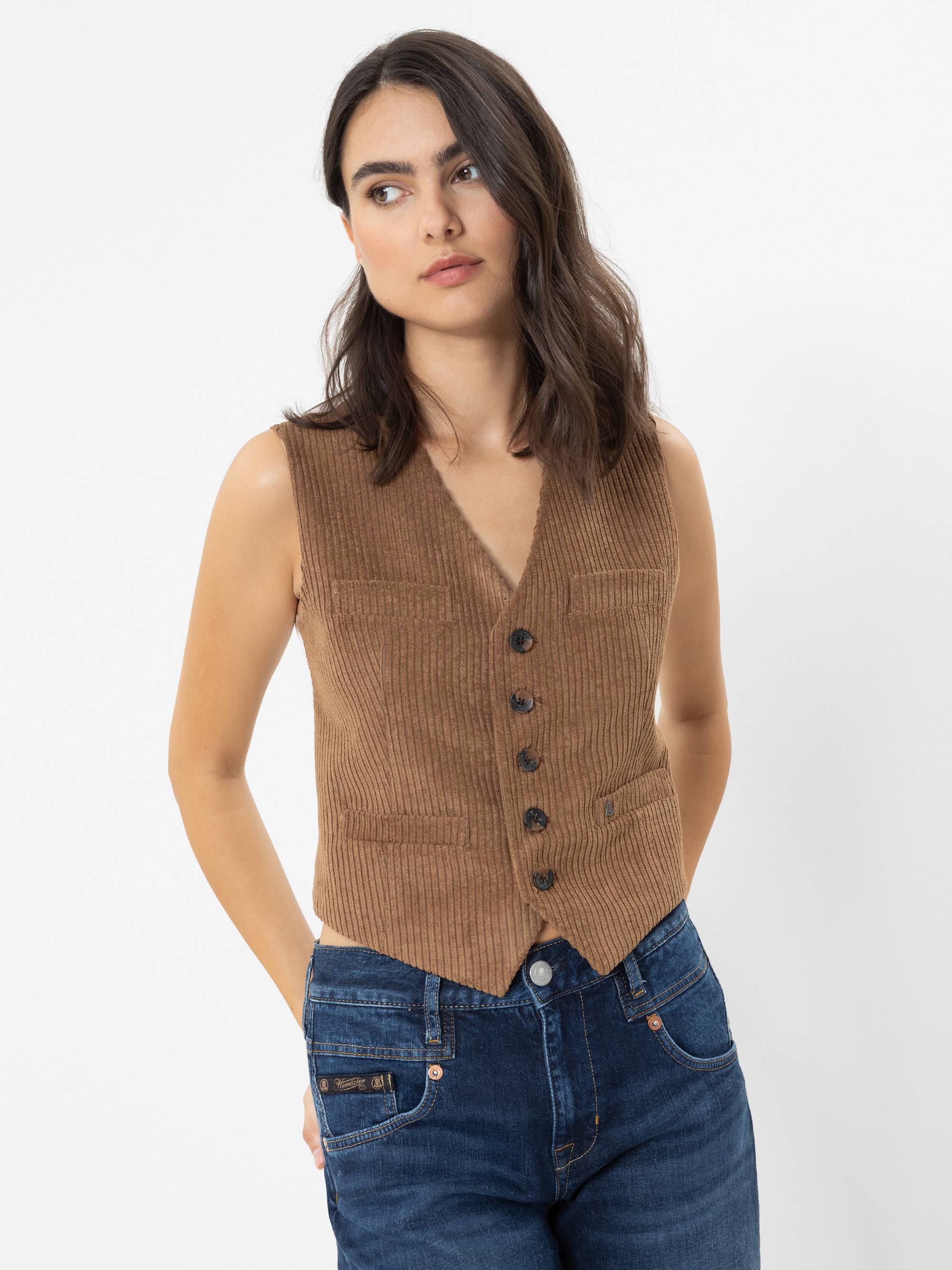 Eine Frau mit welligem braunem Haar trägt eine braune, geknöpfte Weste und blaue Jeans. Sie steht vor einem schlichten weißen Hintergrund, die Hände hinter dem Rücken verschränkt, und blickt mit ausdruckslosem Gesicht leicht zur Seite.