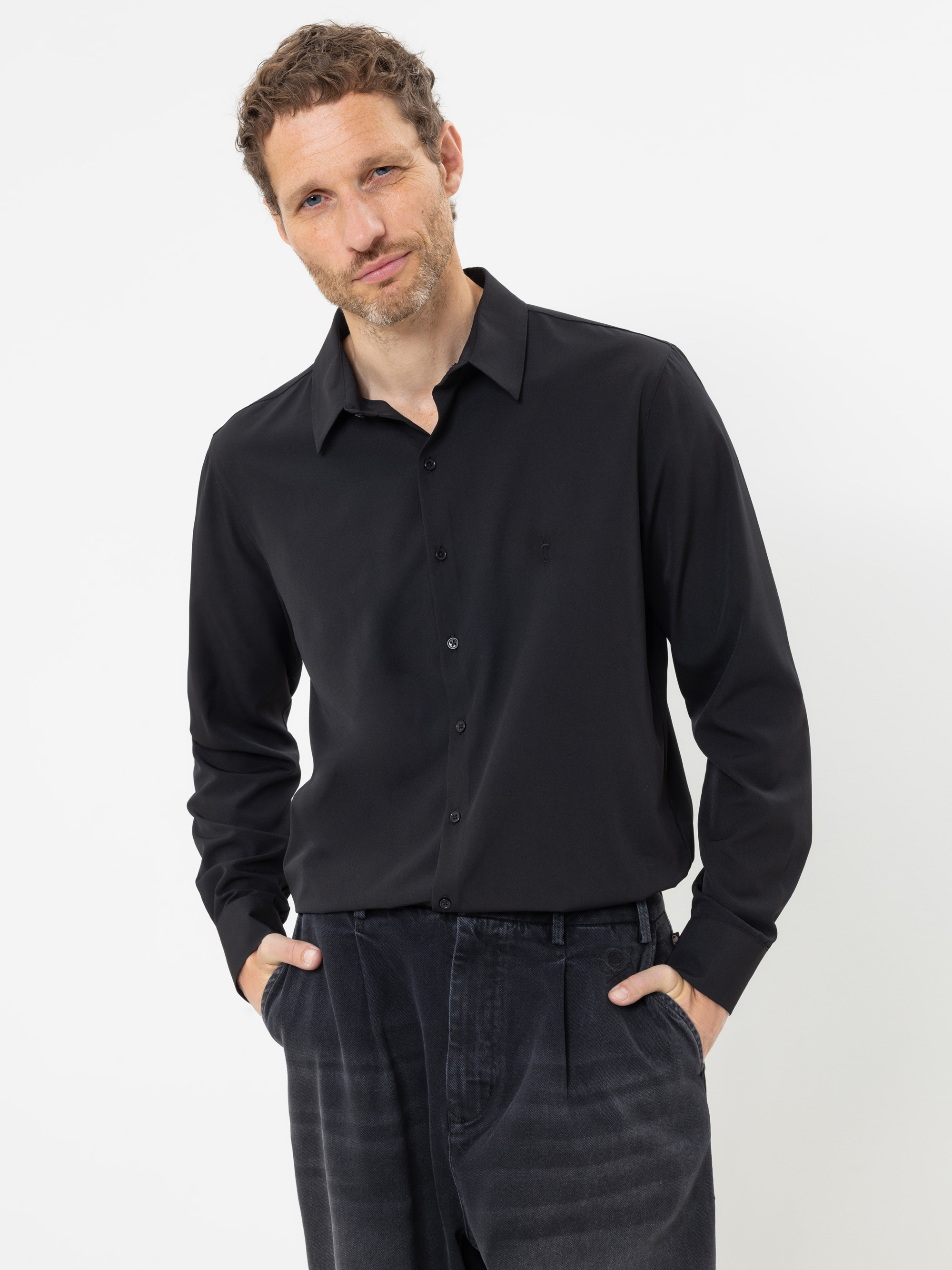 Ein Mann mit kurzen Locken und Bart trägt ein schwarzes Hemd mit Knöpfen und eine dunkle Hose. Er steht vor einem schlichten weißen Hintergrund, die Hände in den Taschen, blickt leicht zur Seite und lächelt sanft.