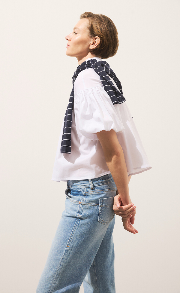 Eine Person in weißer Bluse und hellblauen Jeans steht im Profil mit einem marineblau gestreiften Pullover über den Schultern, die Hände hinter dem Rücken verschränkt, vor einem schlichten hellen Hintergrund.