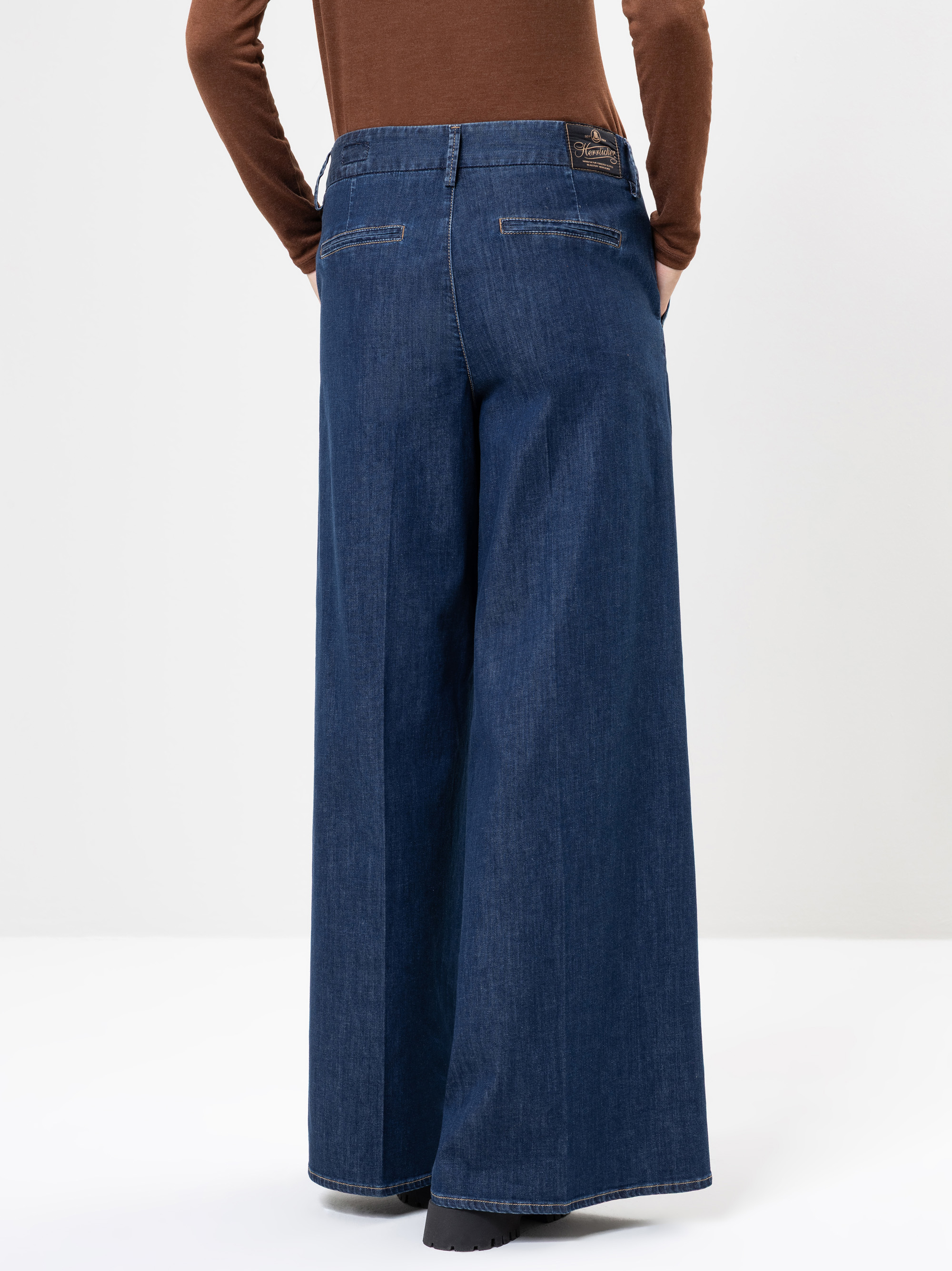 Eine Person, die weite blaue Jeans und ein braunes langärmeliges Oberteil trägt, wird von hinten gezeigt und steht vor einem schlichten weißen Hintergrund.