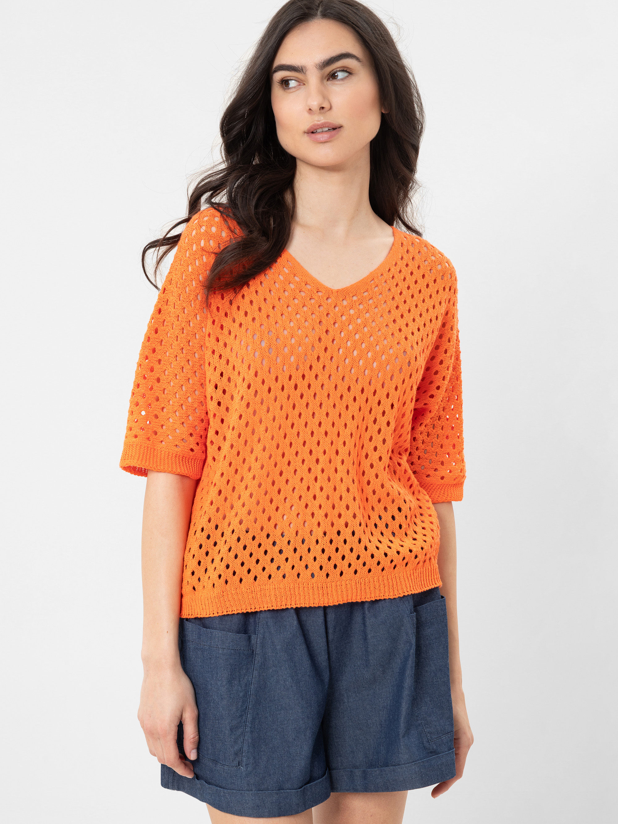 Eine Frau mit langen dunklen Haaren trägt einen leuchtend orangefarbenen, kurzärmeligen, offen gestrickten Pullover mit V-Ausschnitt und blaue Jeans-Shorts und steht vor einem schlichten weißen Hintergrund.