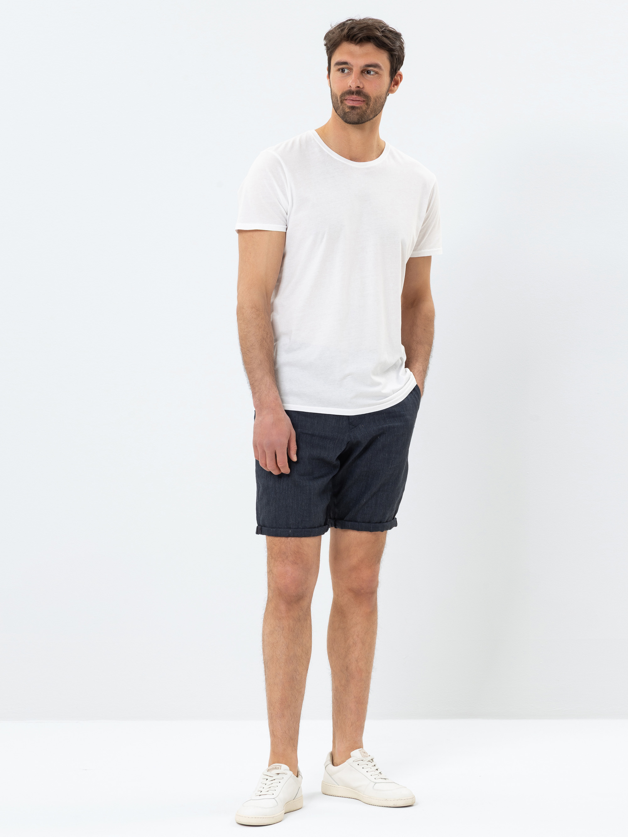 Ein Mann steht vor einem schlichten weißen Hintergrund und trägt ein weißes T-Shirt, dunkelblaue Shorts und weiße Turnschuhe. Er hat kurzes braunes Haar und einen Bart, eine Hand in der Tasche und einen entspannten Gesichtsausdruck.