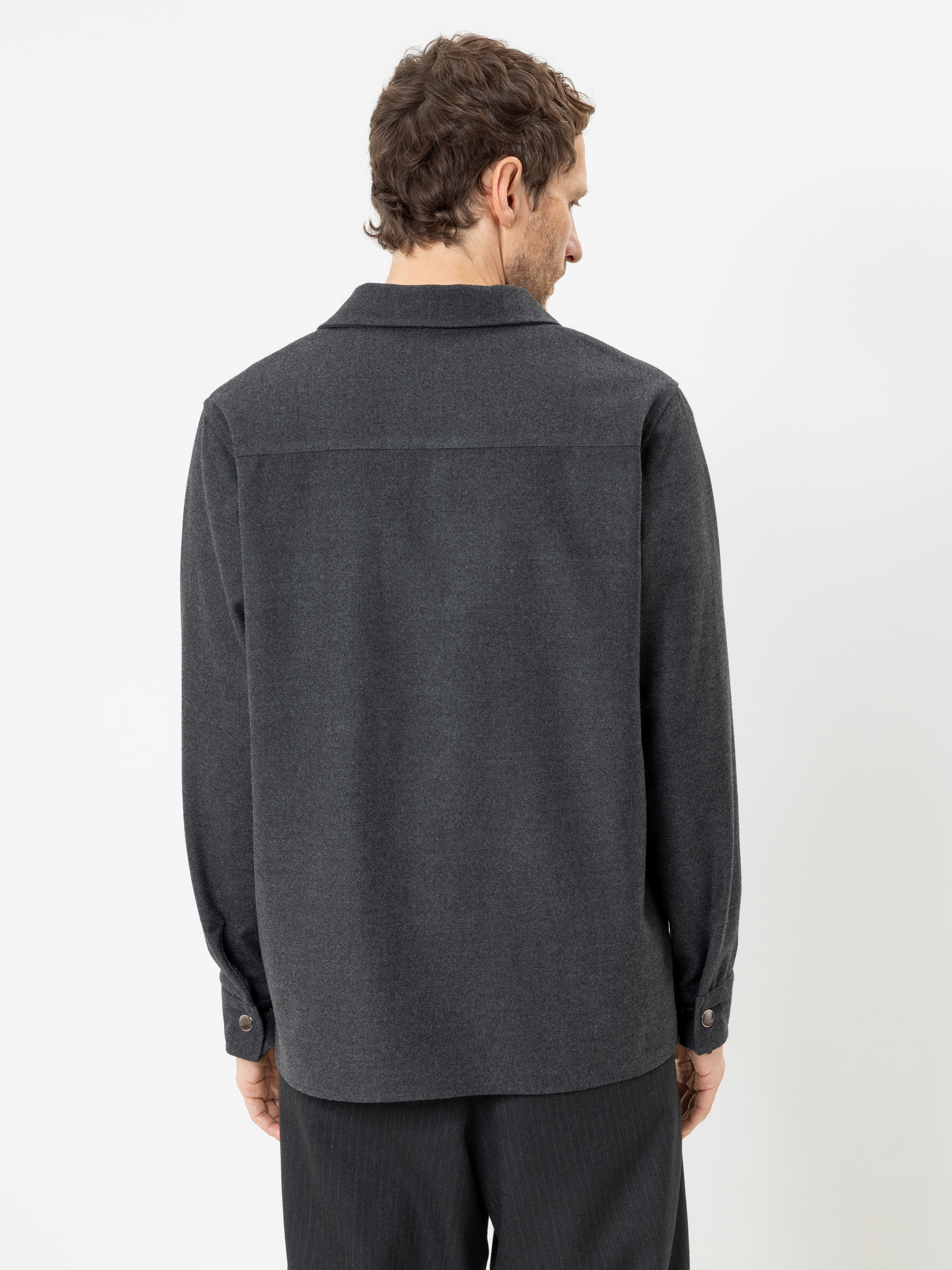 Ein Mann mit kurzen braunen Haaren steht vor einem weißen Hintergrund und ist mit dem Rücken zur Kamera gewandt. Er trägt ein dunkelgraues Langarmhemd und eine schwarze Hose.