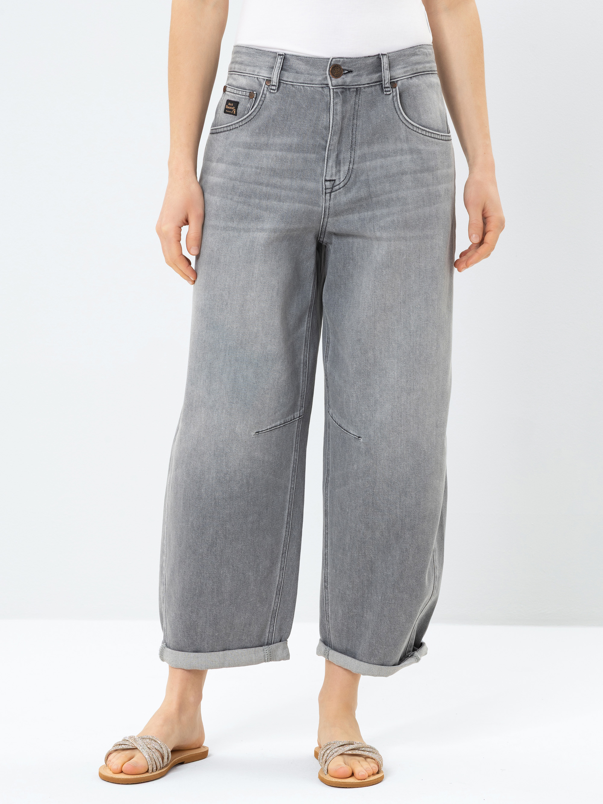 Eine Person, die ein weißes Oberteil, eine locker sitzende graue Jeans mit aufgerollten Bündchen und beigefarbene Sandalen trägt, steht vor einem schlichten weißen Hintergrund. Die Jeans hat sichtbare Nähte und einen lockeren, lässigen Stil.