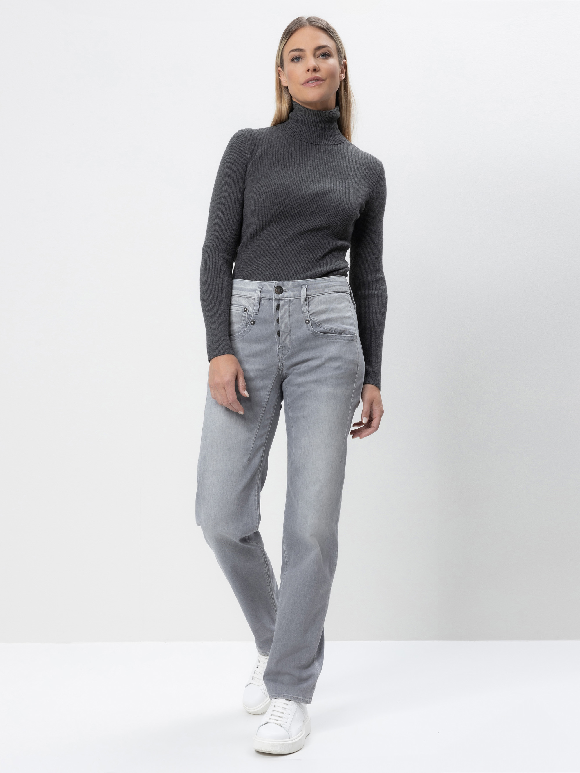 Eine Frau steht vor einem schlichten weißen Hintergrund und trägt einen dunkelgrauen Rollkragenpullover, hellgraue Jeans mit hoher Taille und weiße Turnschuhe. Sie hat glattes hellbraunes Haar und einen neutralen Gesichtsausdruck.