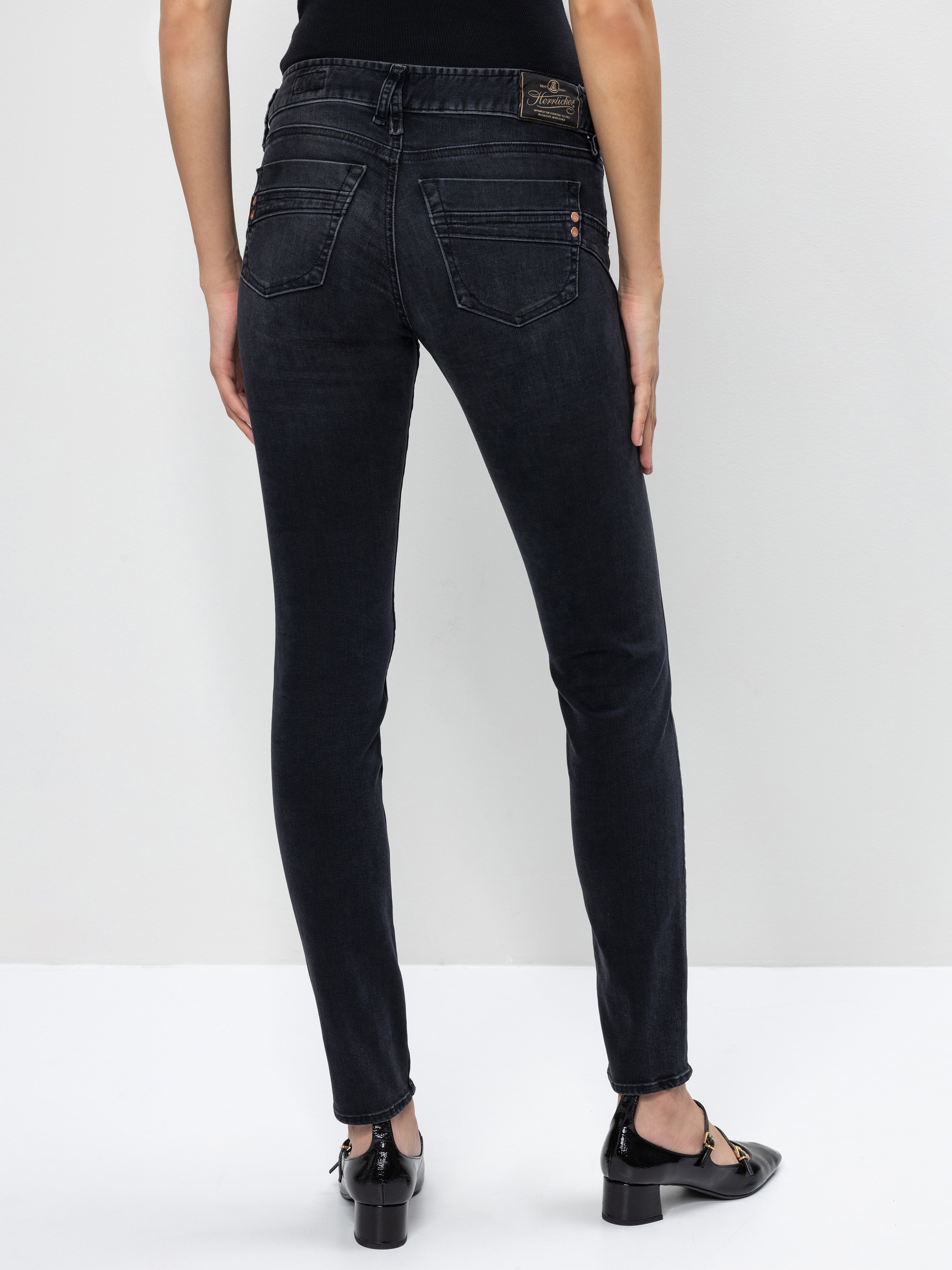 Eine Person, die dunkle Skinny Jeans und schwarze Absätze trägt, steht mit dem Rücken zur Kamera vor einem schlichten hellen Hintergrund. Ihre Arme liegen entspannt an der Seite.