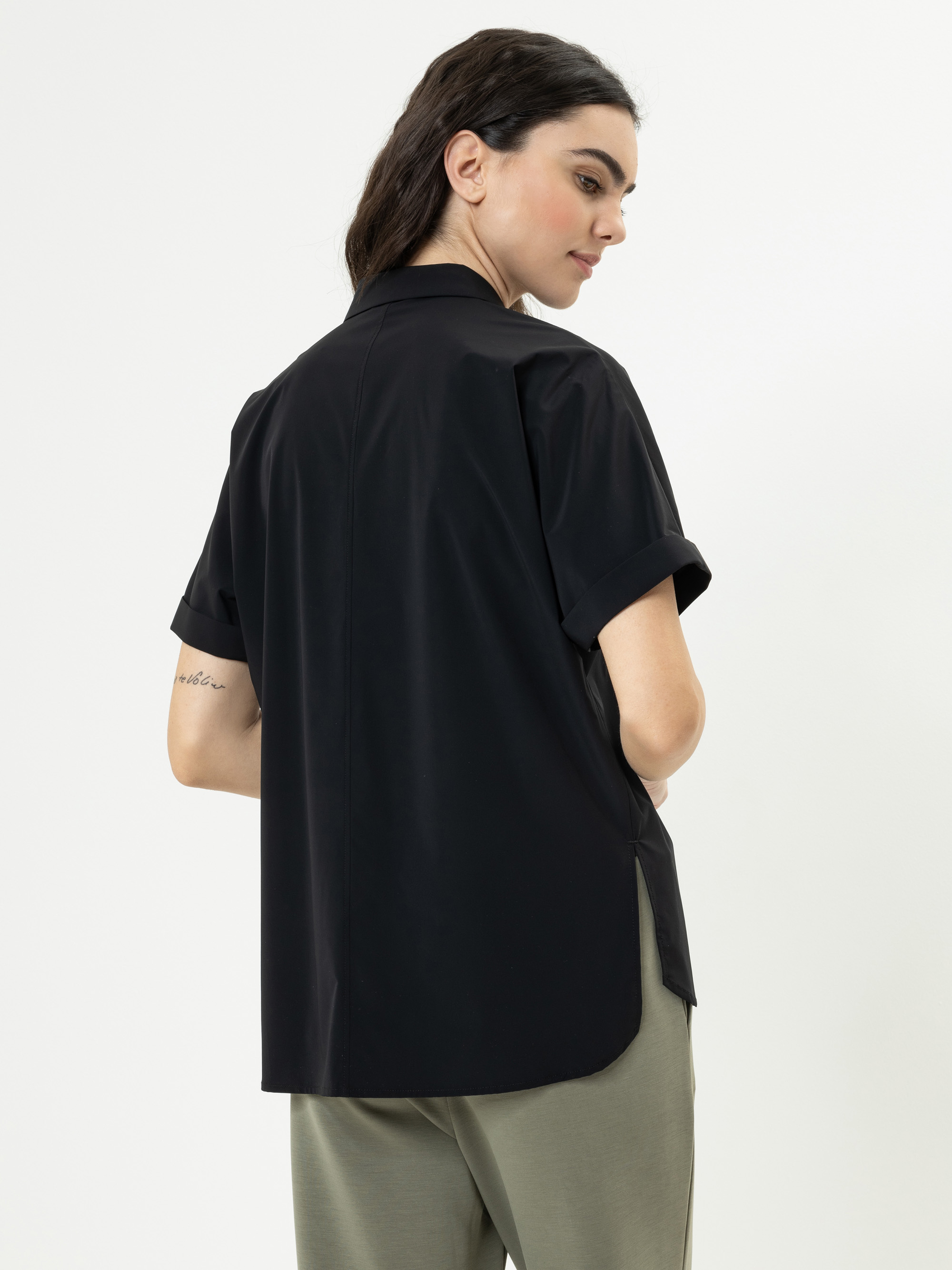 Eine Frau mit langen dunklen Haaren, bekleidet mit einem schwarzen kurzärmeligen Button-up-Hemd und einer hellen olivgrünen Hose, steht mit dem Rücken zur Kamera und schaut über ihre Schulter. Auf ihrem rechten Unterarm ist eine kleine Tätowierung zu