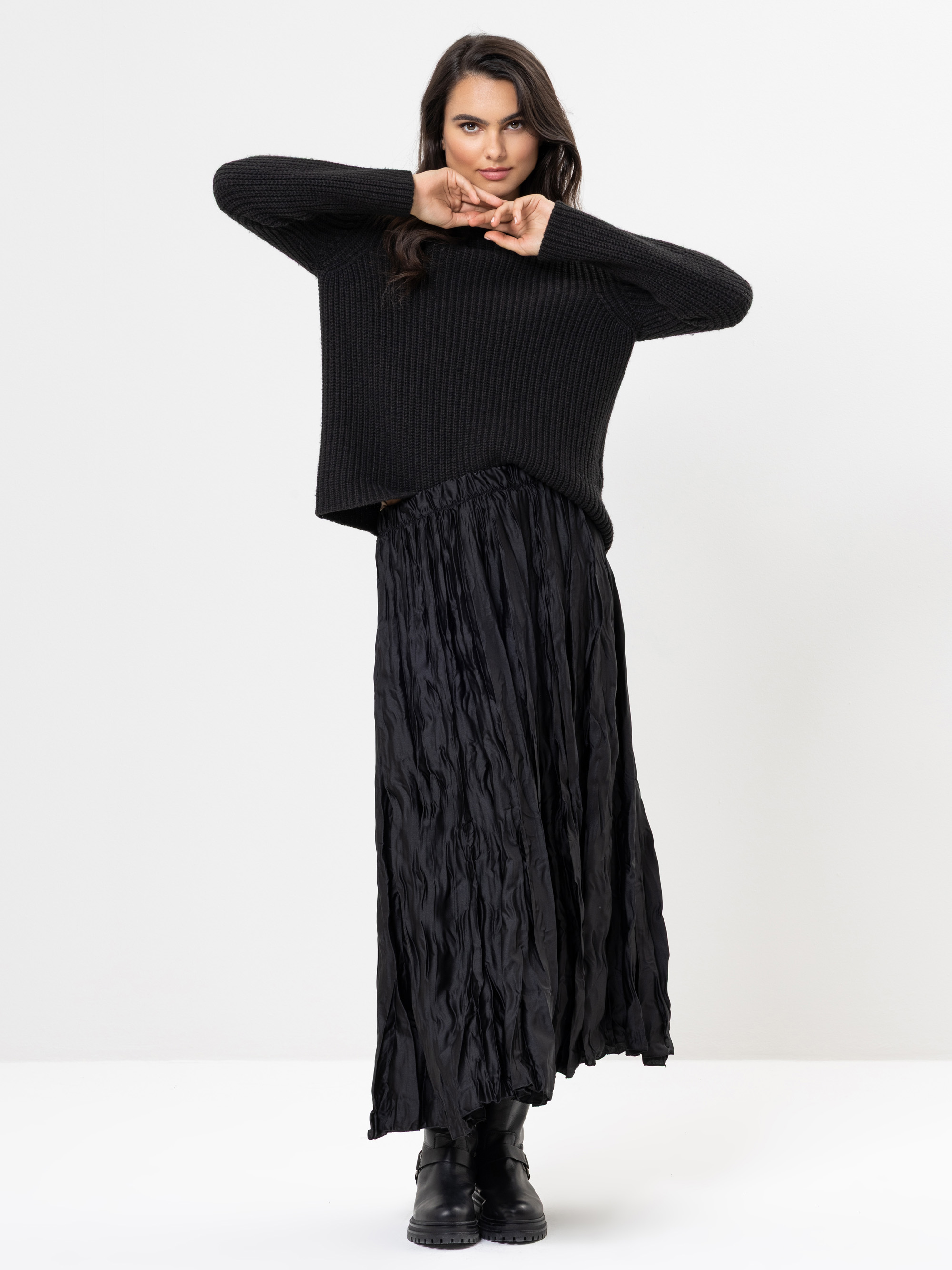Eine Frau steht vor einem weißen Hintergrund und trägt einen lockeren schwarzen Pullover, einen langen, zerknitterten schwarzen Rock und schwarze Stiefel. Sie hat dunkles Haar und posiert mit angewinkelten Armen, die Hände vor dem Gesicht und einem sel