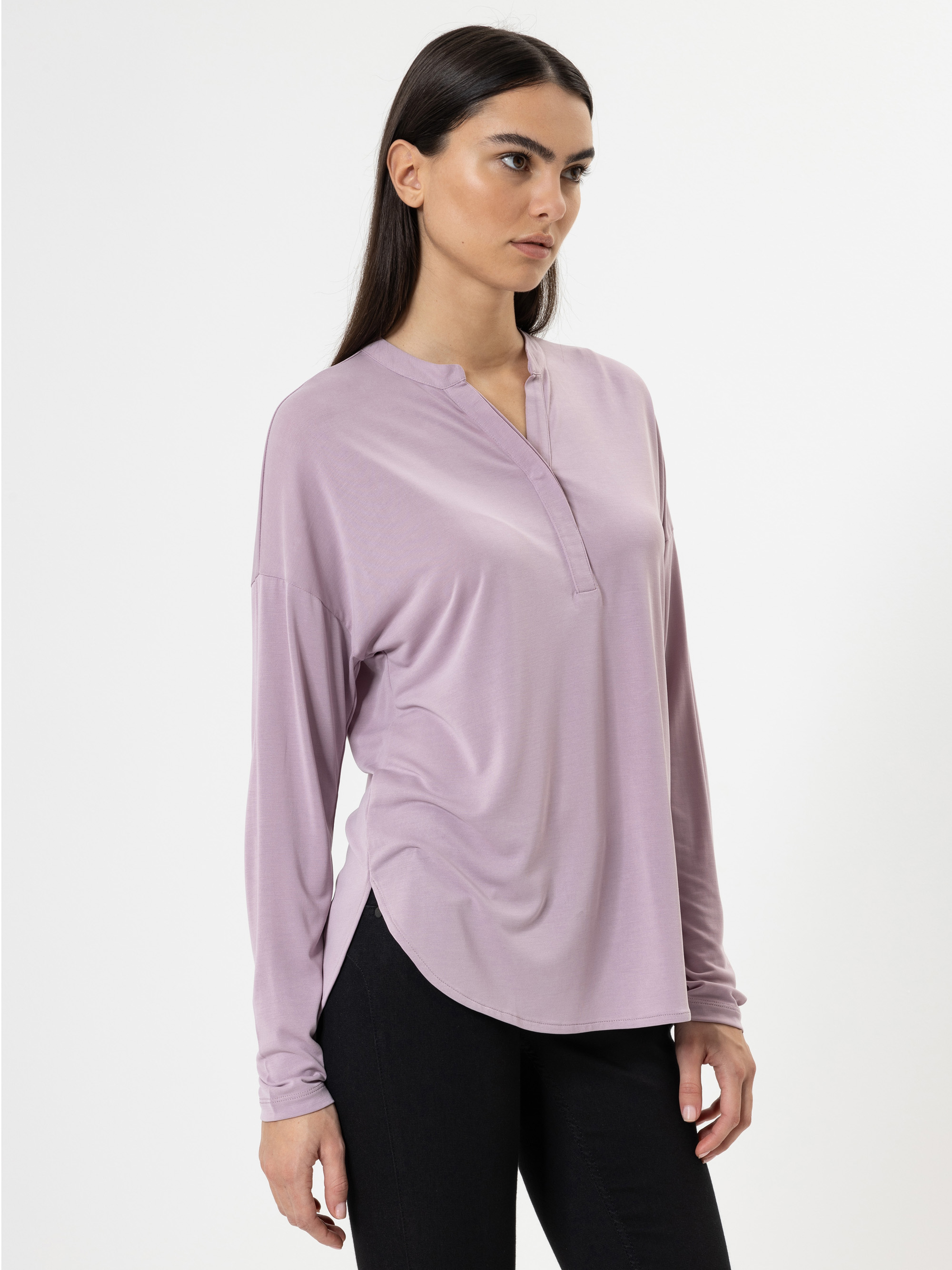 Eine Frau mit langen dunklen Haaren, die eine locker sitzende, hellviolette, langärmelige Bluse und eine schwarze Hose trägt, steht vor einem schlichten weißen Hintergrund und schaut leicht zur Seite.