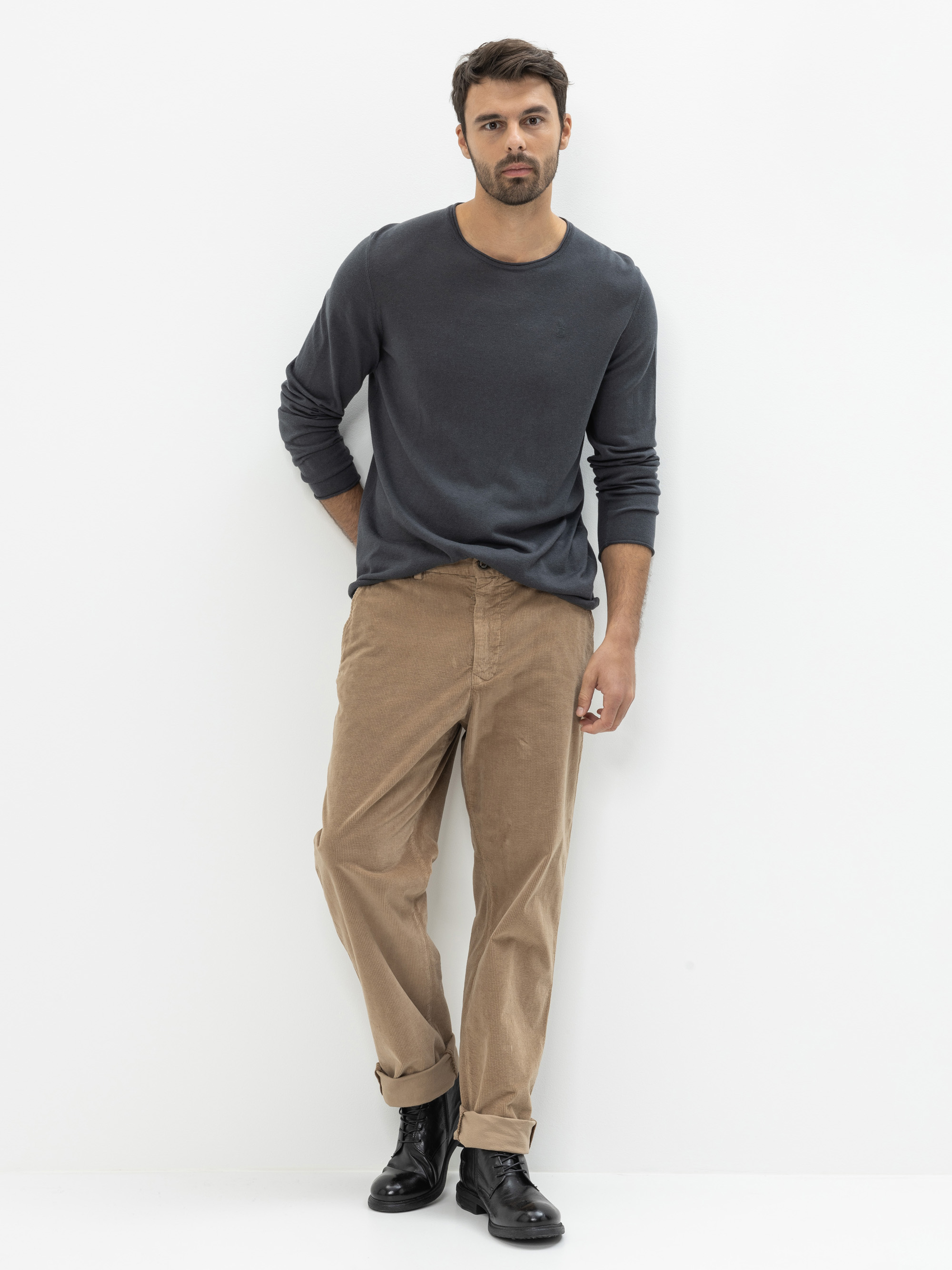 Ein Mann steht vor einem weißen Hintergrund und trägt ein langärmeliges dunkelgraues Hemd, eine weite hellbraune Hose mit Manschetten und schwarze Schnürstiefel. Er hat einen kurzen Bart und dunkles Haar, seine Pose und sein Gesichtsausdruck sind ents