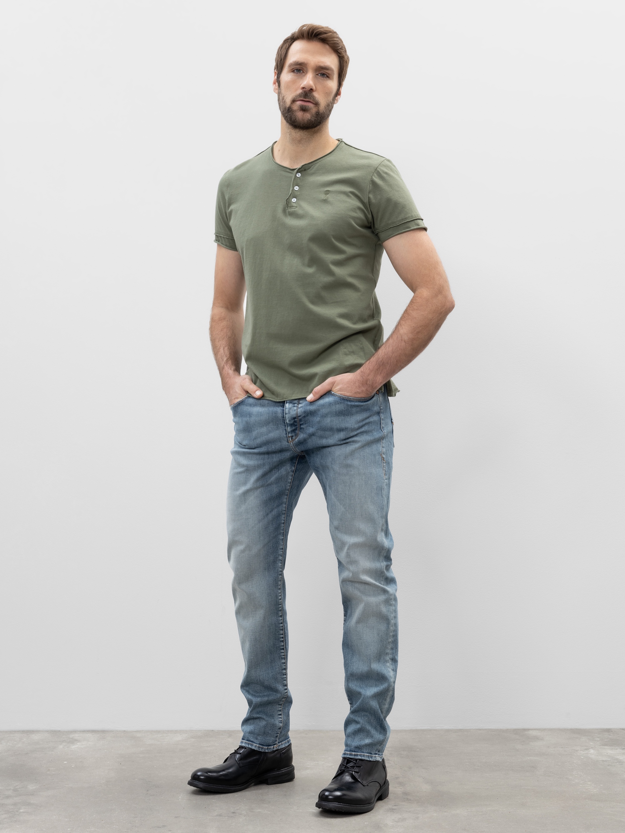 Ein Mann mit kurzen braunen Haaren und einem Bart steht vor einem schlichten weißen Hintergrund. Er trägt ein grünes, kurzärmeliges Hemd, hellblaue Jeans und schwarze Schuhe, die Hände in den Taschen.