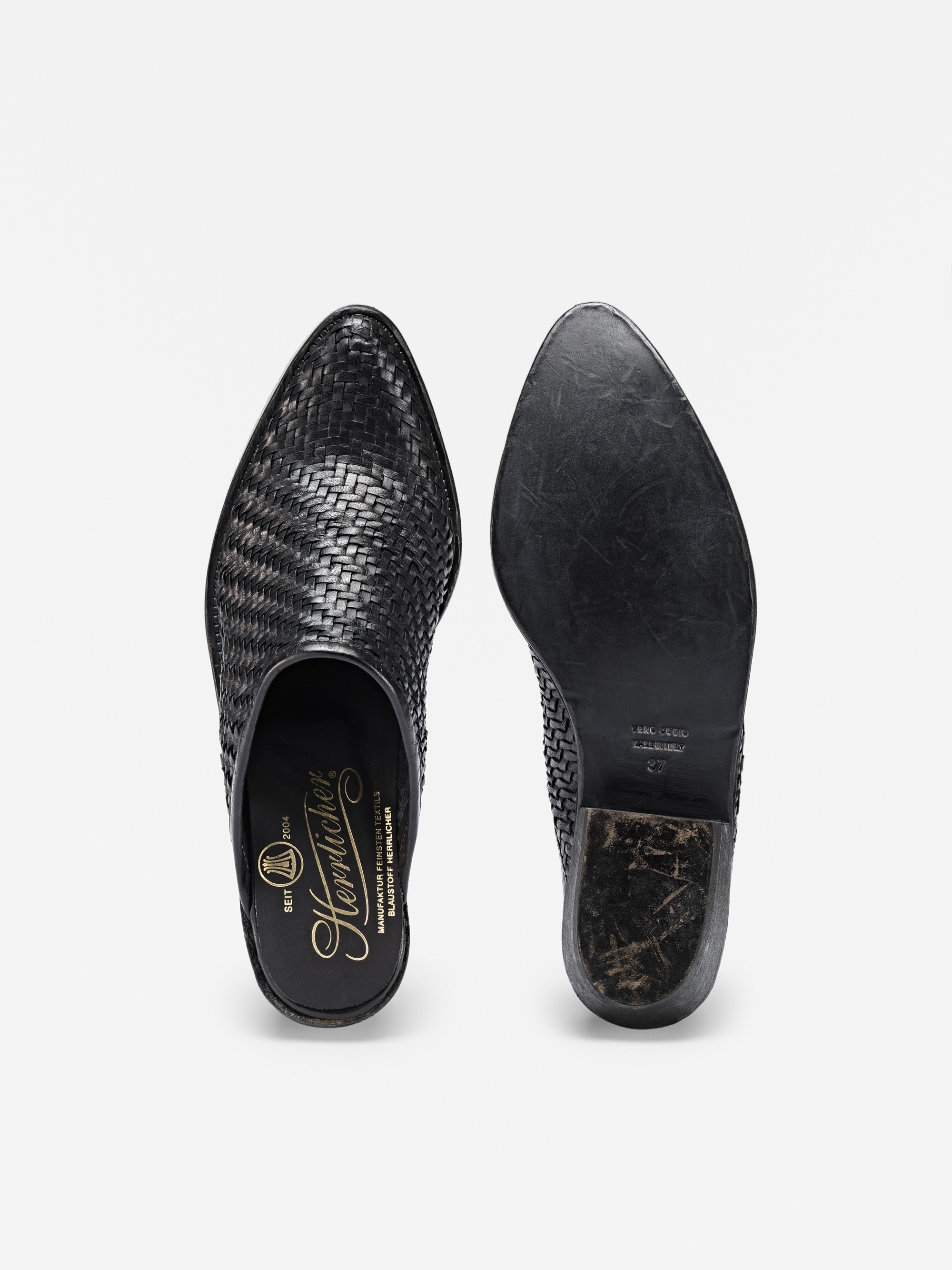 Ein Paar schwarzer Slipper aus gewebtem Leder von oben; der linke Schuh zeigt das Branding auf dem Oberteil und der Innensohle, während der rechte Schuh die abgenutzte Sohle zeigt.