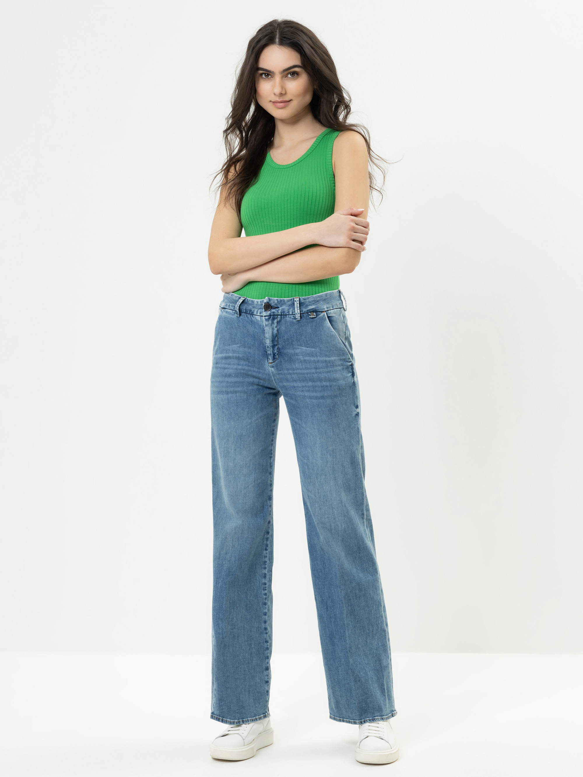 Eine Frau mit langen dunklen Haaren, die ein grünes ärmelloses Oberteil, blaue Jeans mit hoher Taille und weiße Turnschuhe trägt, steht selbstbewusst mit verschränkten Armen in einem Studio mit einem schlichten weißen Hintergrund.