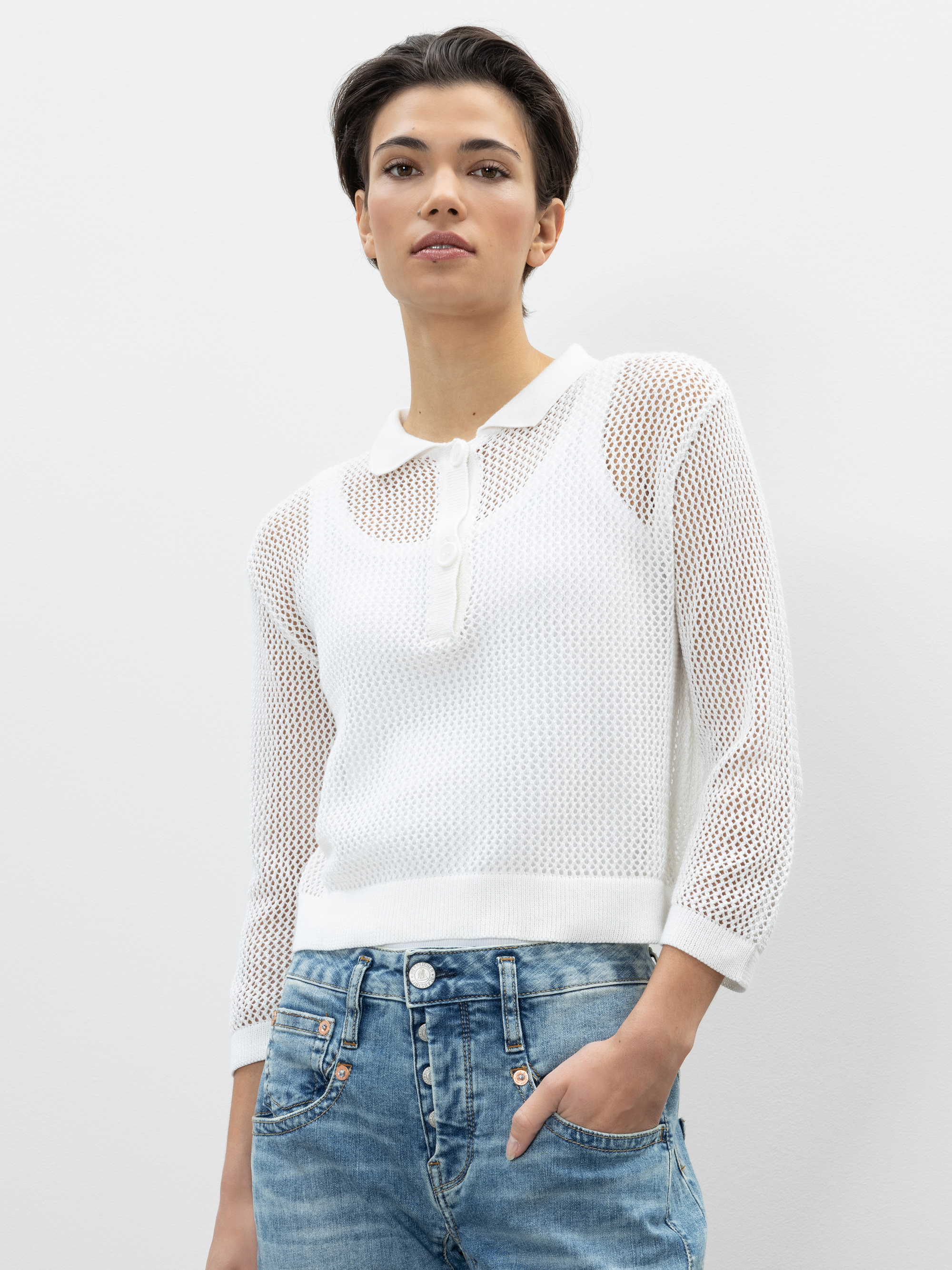 Eine Person, die einen durchsichtigen weißen langärmeligen Strickpullover mit Kragen und Knöpfen und eine hellblaue Jeans trägt, steht vor einem einfarbig weißen Hintergrund.