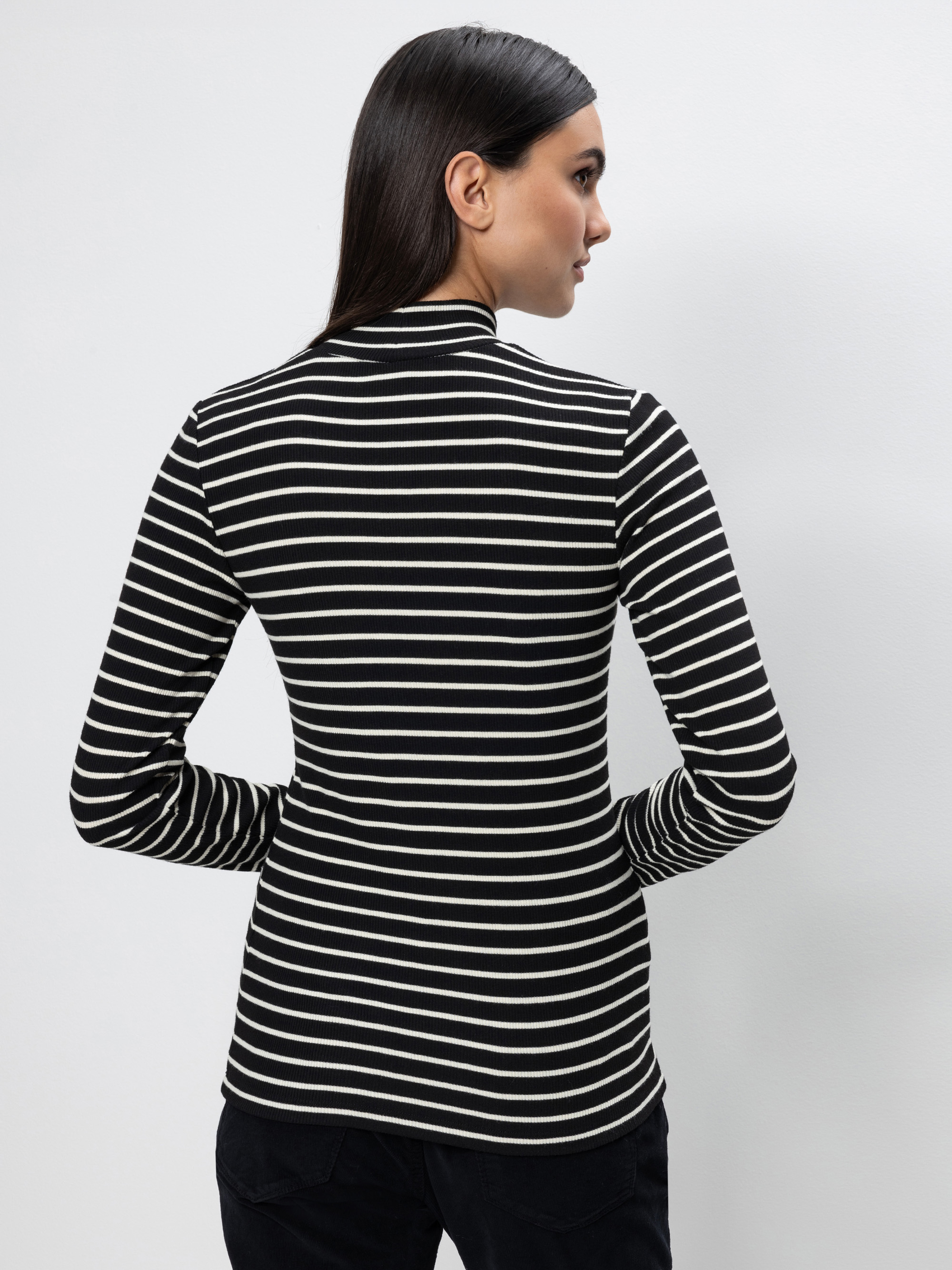 Eine Frau mit glattem dunklem Haar steht mit dem Gesicht zur Wand. Sie trägt ein schwarz-weißes, langärmeliges, horizontal gestreiftes Oberteil mit hohem Ausschnitt und eine schwarze Hose vor einem schlichten hellgrauen Hintergrund.