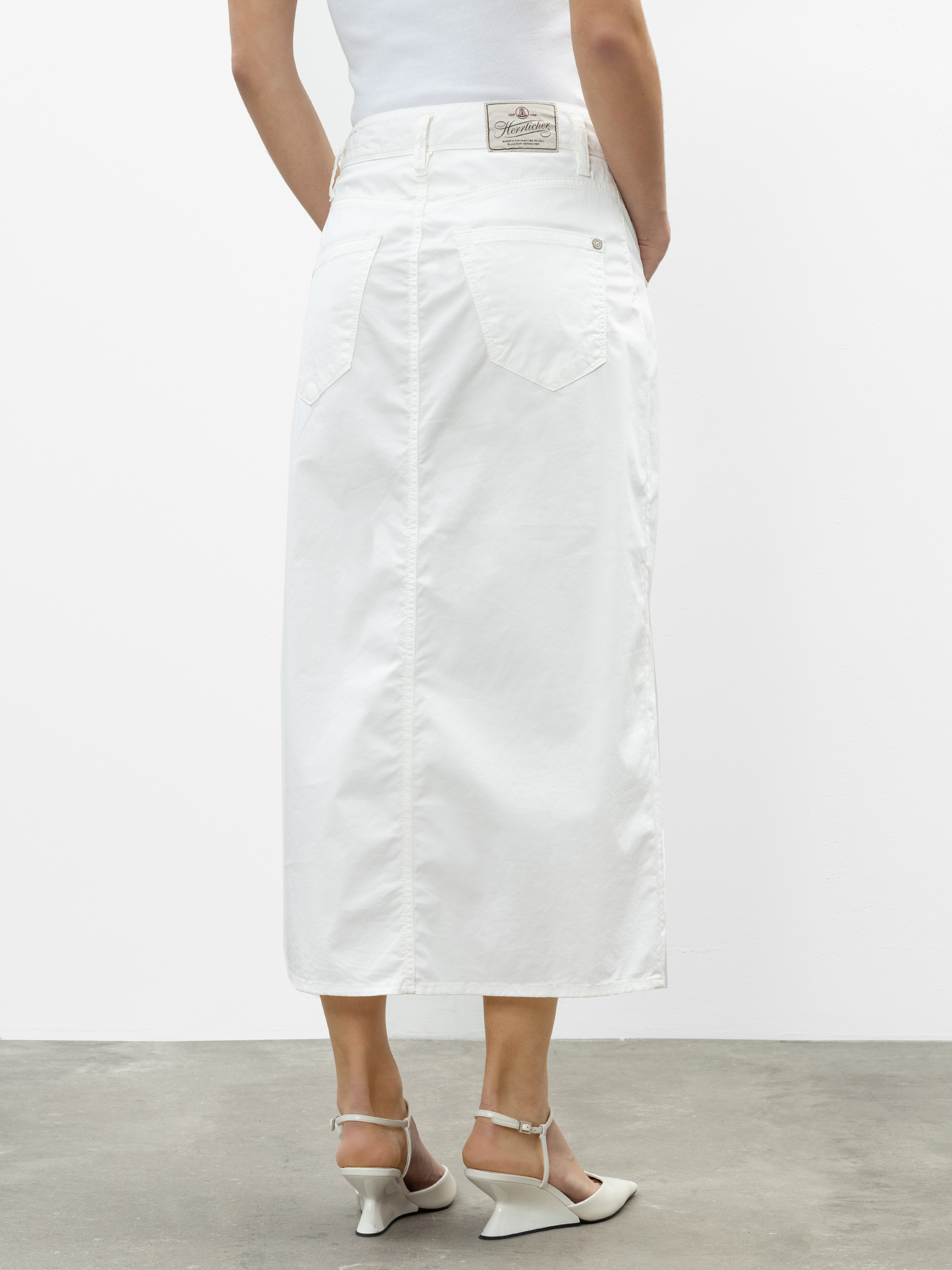 Eine Person, die einen langen weißen Jeansrock mit Gesäßtaschen und einem Etikett trägt, dazu ein weißes ärmelloses Oberteil und weiße hochhackige Sandalen, steht auf einem Betonboden und schaut von der Kamera weg.