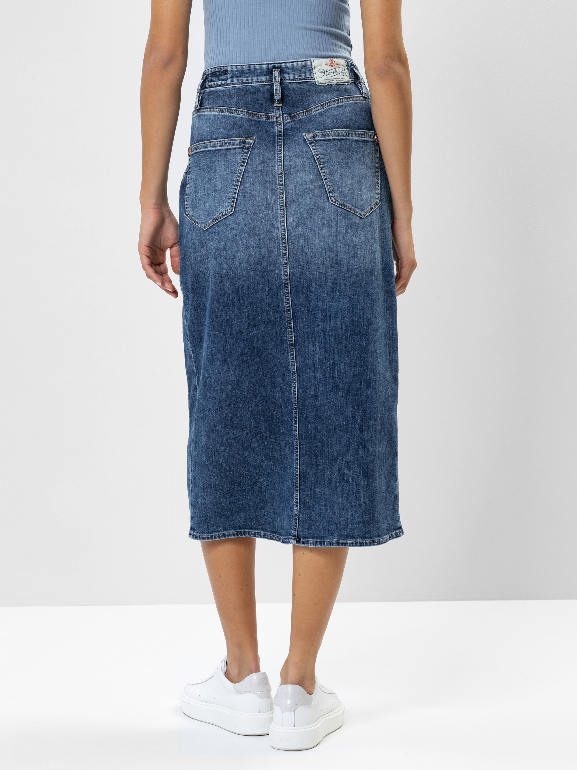 Eine Person, die einen blauen Jeans-Midirock und weiße Turnschuhe trägt, steht mit dem Gesicht zur Seite und zeigt die Rückseite des Rocks mit zwei Taschen und verblassten Details. Der Oberkörper ist mit einem hellblauen Oberteil bekleidet.