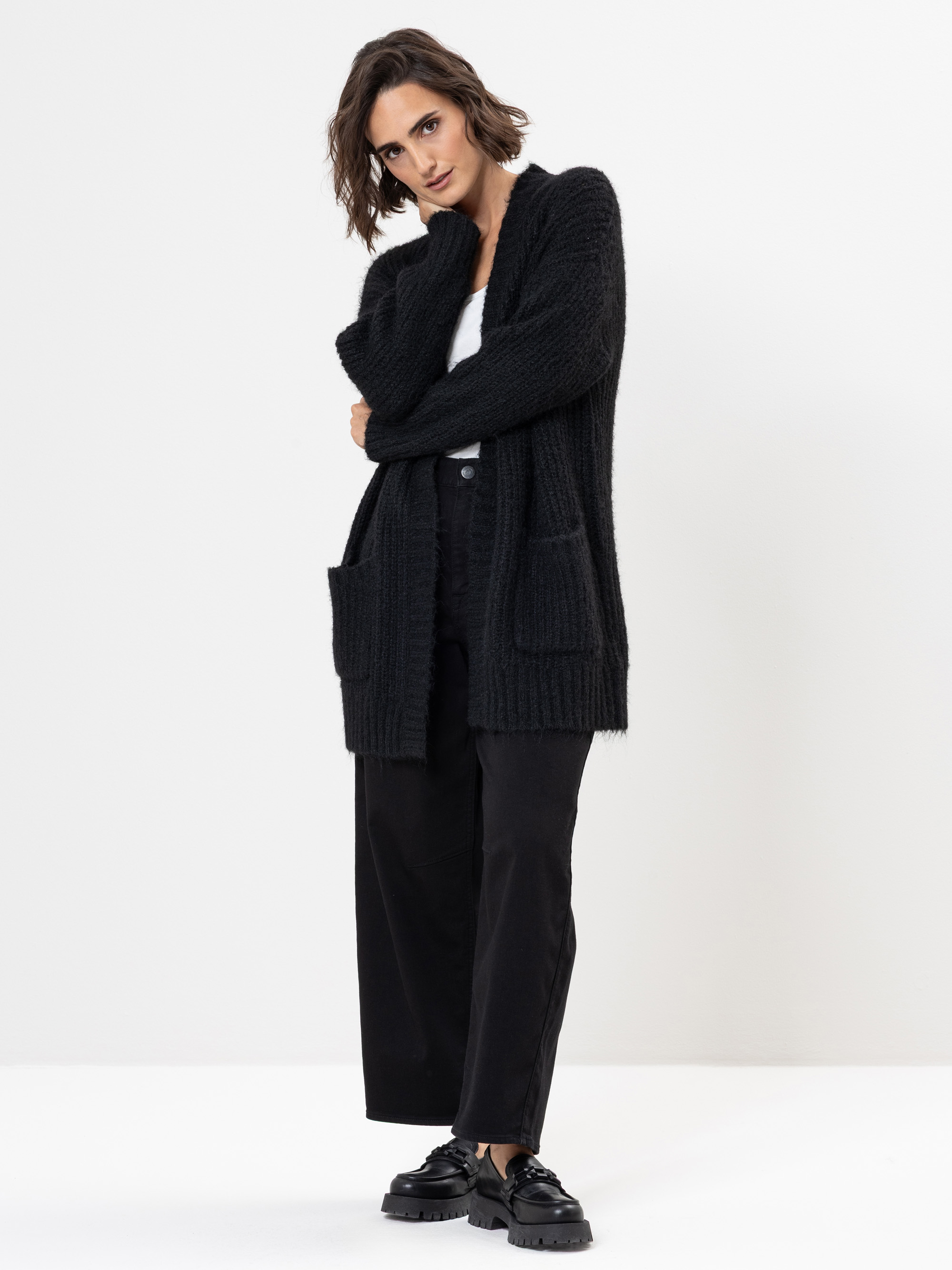 Eine Person mit kurzen braunen Haaren trägt eine schwarze, strukturierte Strickjacke mit Vordertaschen, ein weißes Oberteil, eine schwarze Hose mit weitem Bein und schwarze, klobige Schuhe vor einem schlichten weißen Hintergrund.