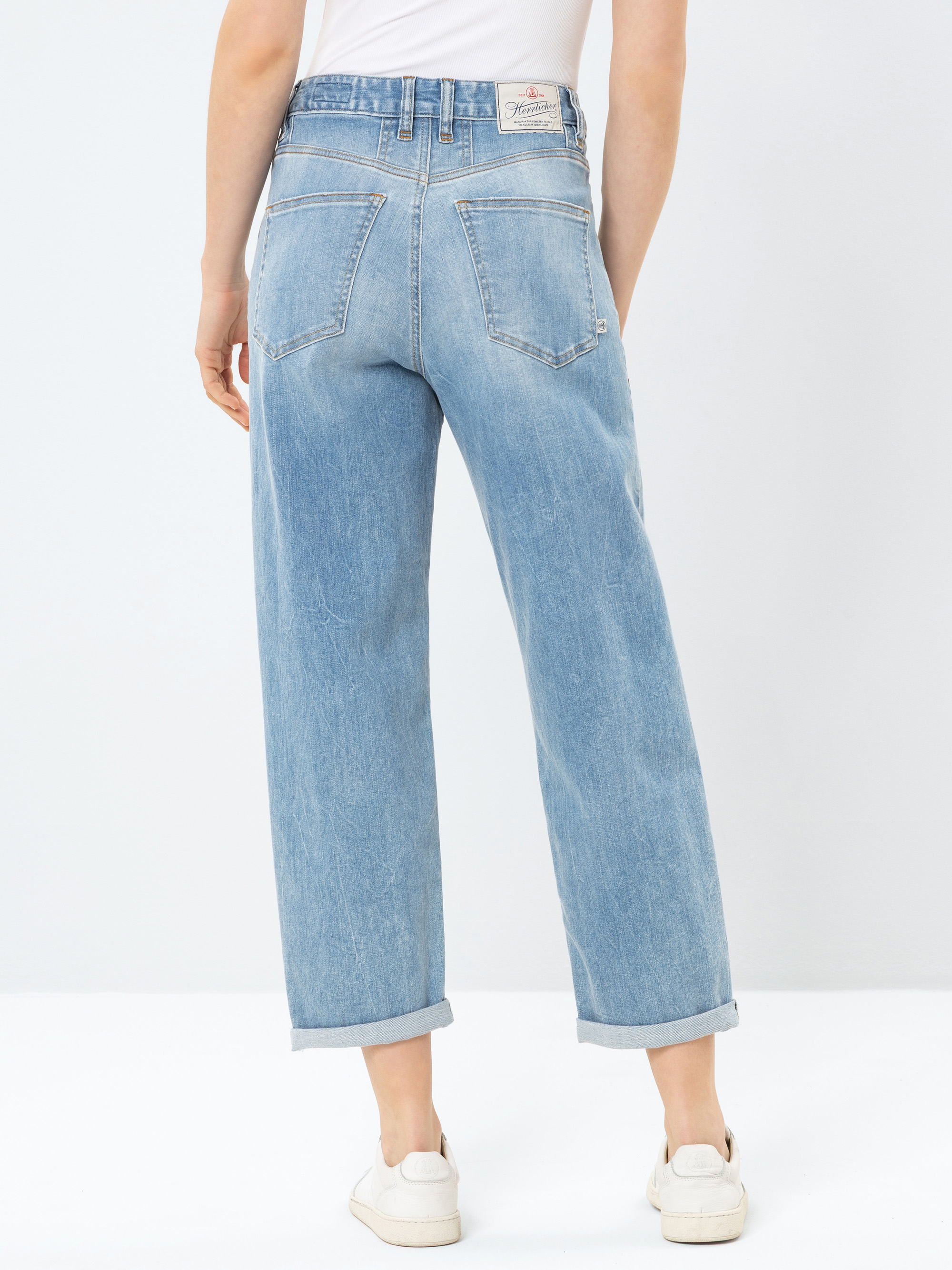 Person, die eine hellblaue Jeans mit hoher Taille und geradem Bein mit gekrempelten Bündchen, ein weißes Oberteil und weiße Turnschuhe trägt. Die Abbildung zeigt die Rückansicht, auf der zwei Gesäßtaschen und ein Markenetikett am Bund zu sehen sind
