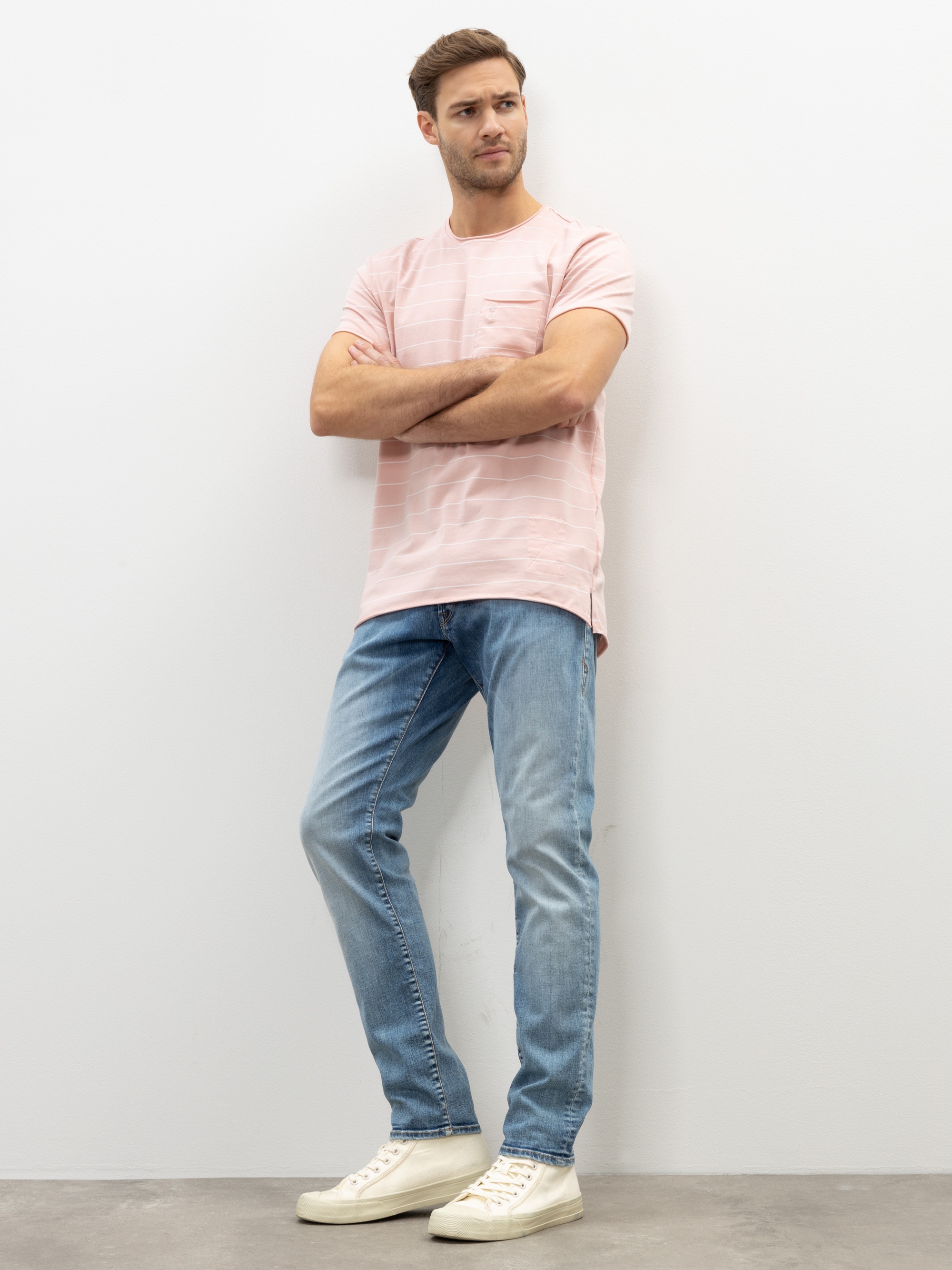 Ein Mann, der ein hellrosa kurzärmeliges T-Shirt, hellblaue Jeans und weiße Turnschuhe trägt, steht mit verschränkten Armen vor einer weißen Wand und schaut leicht zur Seite.