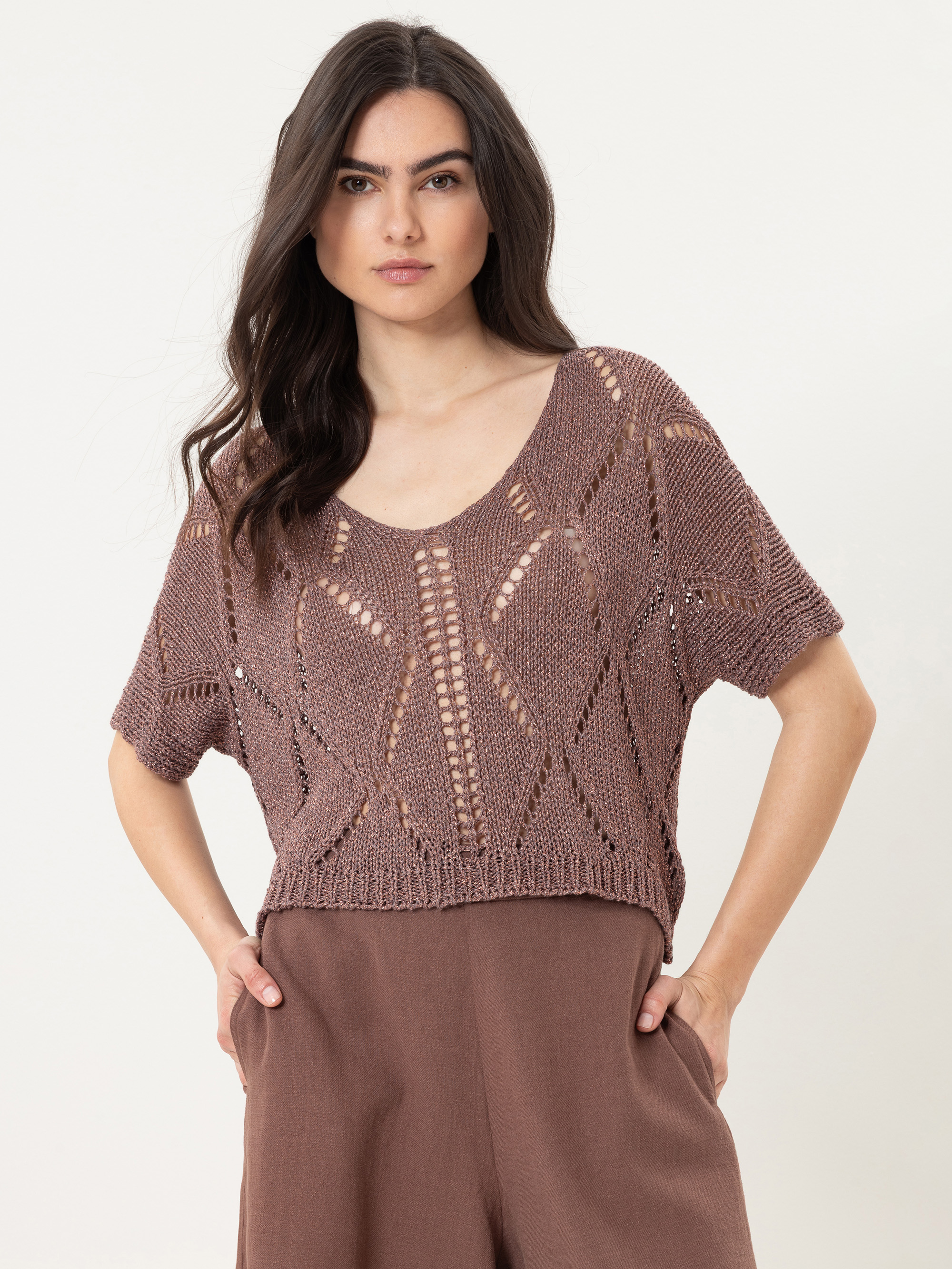 Eine Frau mit langen dunklen Haaren trägt ein lilafarbenes, gehäkeltes, kurzärmeliges Strickoberteil mit Rundhalsausschnitt und eine braune Hose mit hoher Taille. Sie steht mit den Händen in den Taschen vor einem schlichten hellen Hintergrund.
