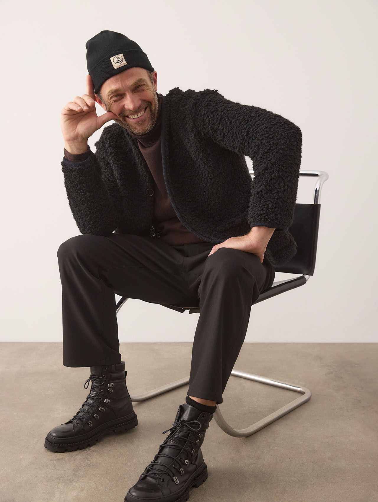 Ein lächelnder Mann, der eine schwarze Mütze, eine strukturierte schwarze Jacke, eine dunkle Hose und schwarze Stiefel trägt, sitzt auf einem modernen Stuhl und berührt mit einer Hand seinen Hut. Der Hintergrund ist schlicht und in hellen Farben gehal