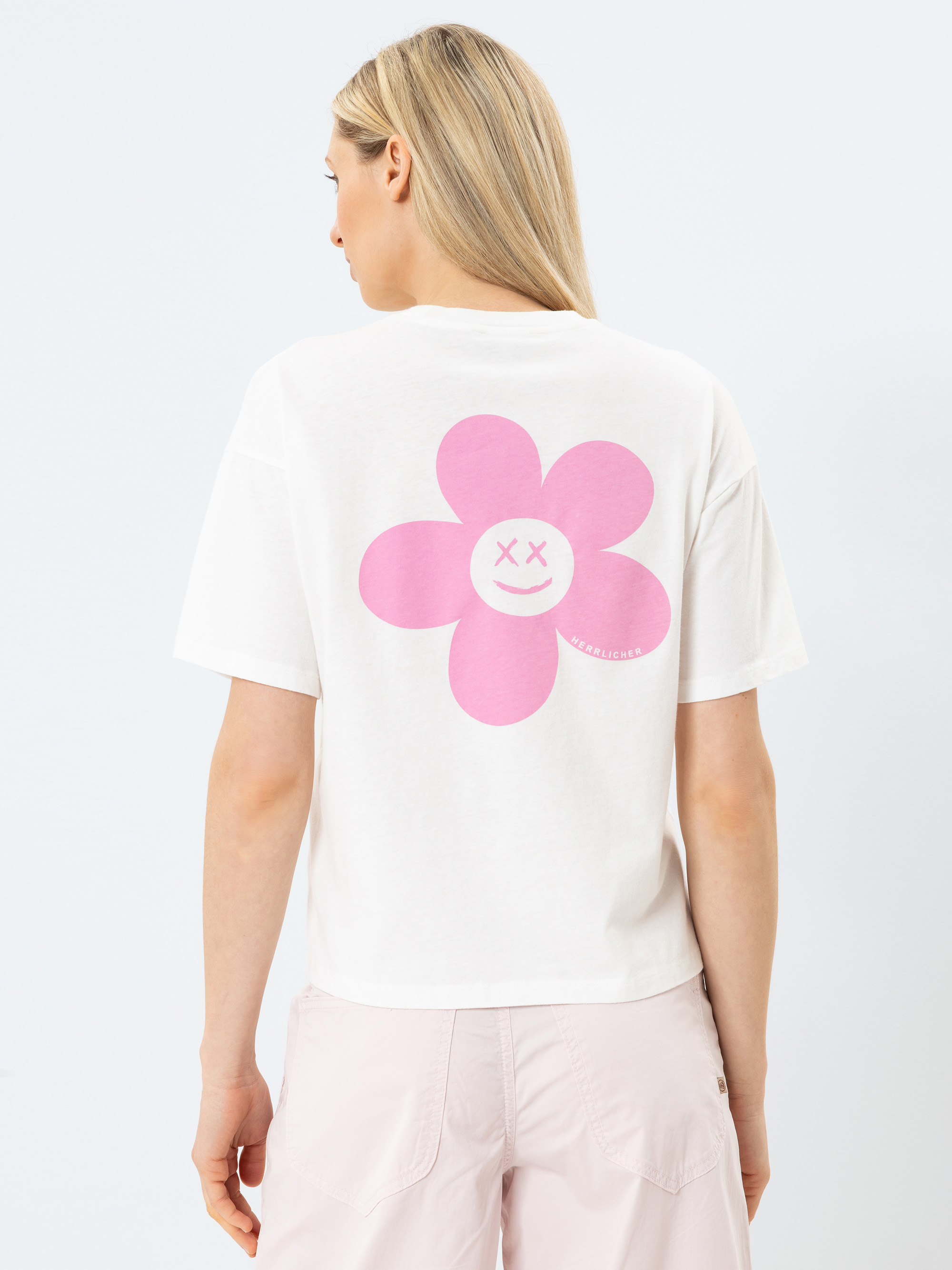 Eine Frau mit langen blonden Haaren trägt ein weißes T-Shirt mit einer großen rosa Blume und einem lächelnden Gesicht mit X-Augen auf dem Rücken. Sie blickt von der Kamera weg, und ihre Shorts sind hellrosa.