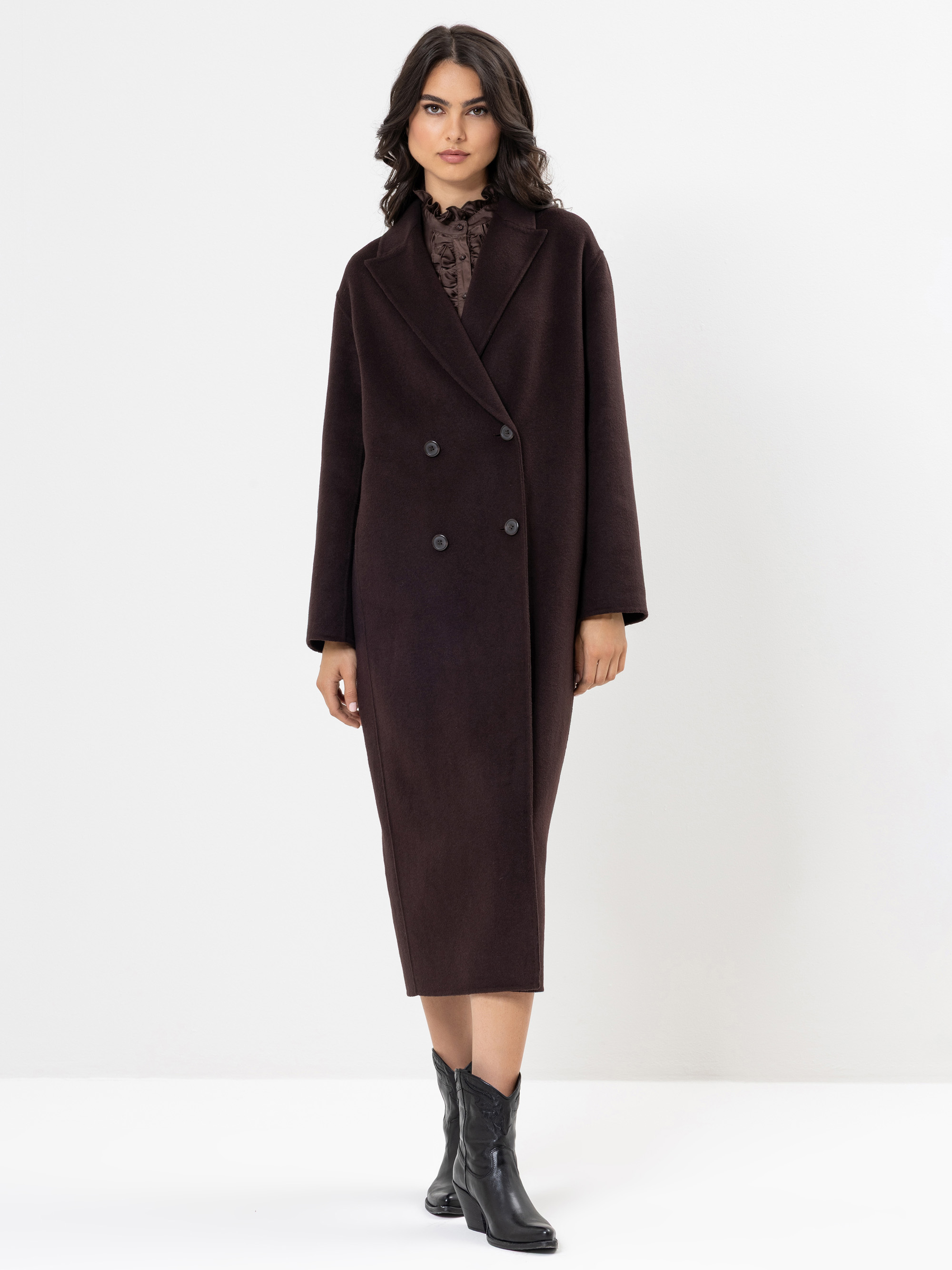 Eine Frau, die einen langen, dunkelbraunen Zweireiher über einem hochgeschlossenen Oberteil und schwarze Ankle Boots trägt, steht vor einem schlichten weißen Hintergrund.