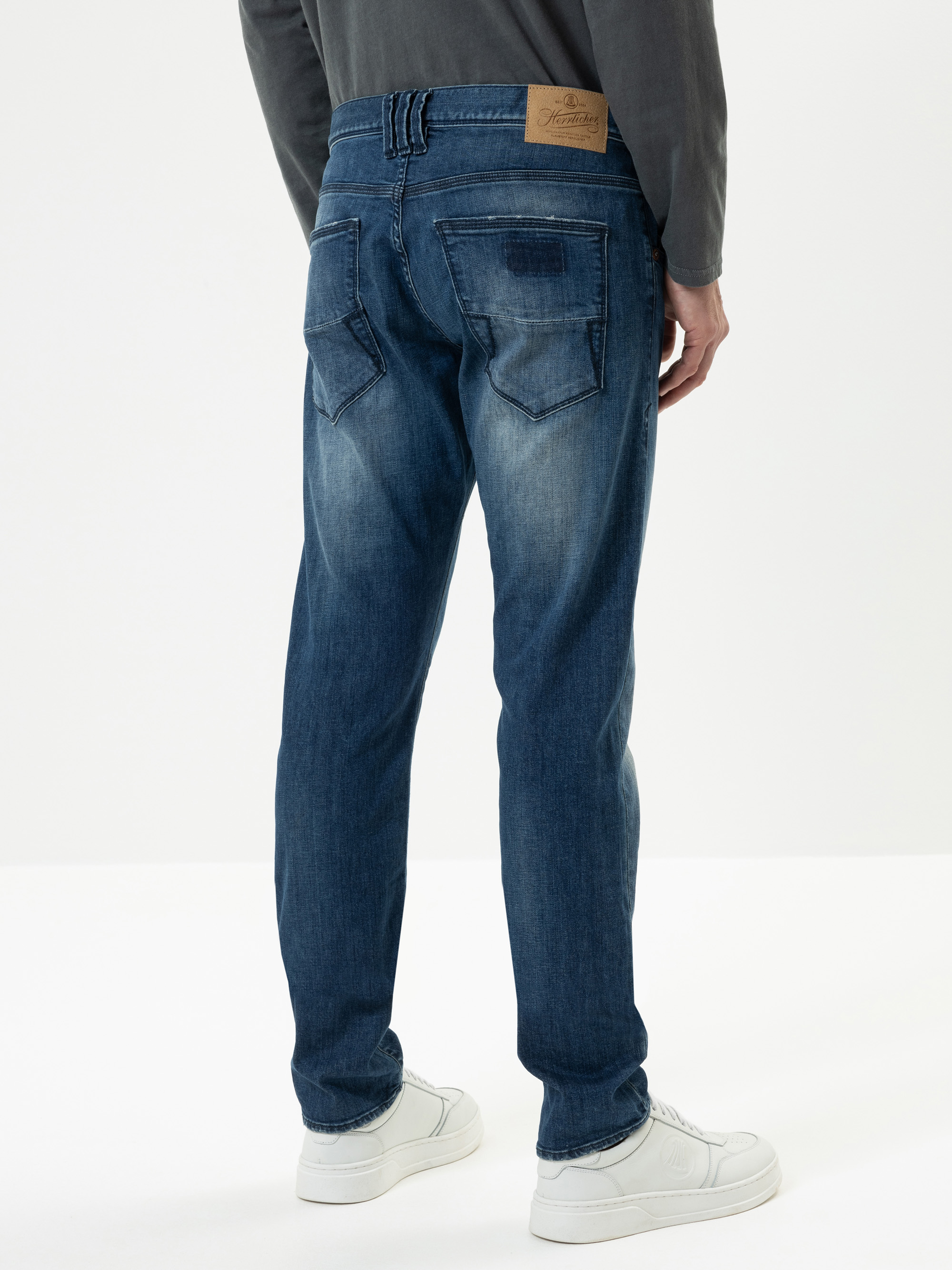 Eine Person, die blaue Jeans und weiße Turnschuhe trägt, wird von hinten gezeigt, vor einem schlichten weißen Hintergrund. Die Jeans hat verblasste Details und einen braunen Lederaufnäher am Bund.