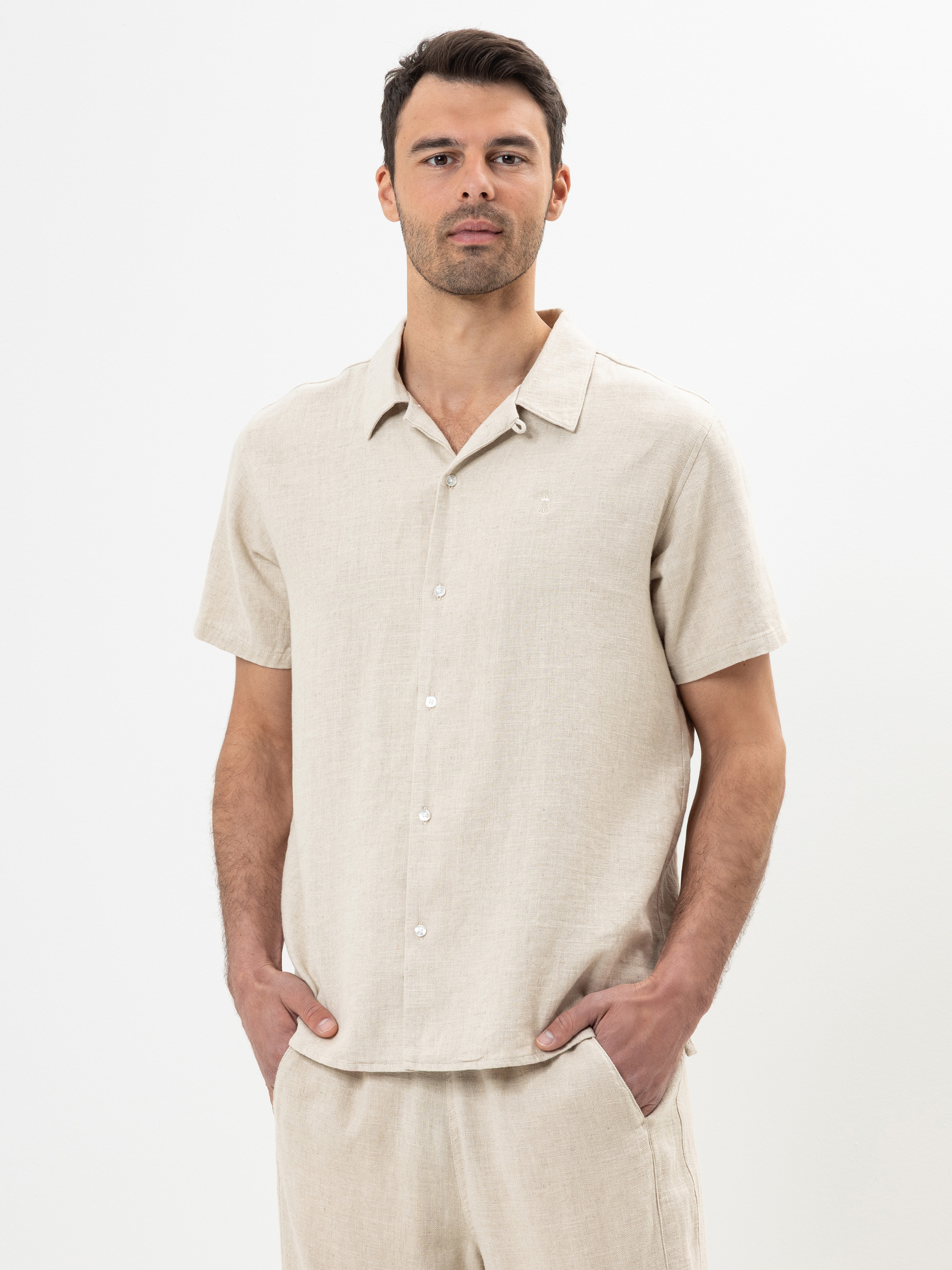 Ein Mann, der ein hellbeiges, kurzärmeliges Hemd mit Knopfleiste und eine dazu passende beigefarbene Hose trägt, steht mit den Händen in den Taschen vor einem schlichten weißen Hintergrund.