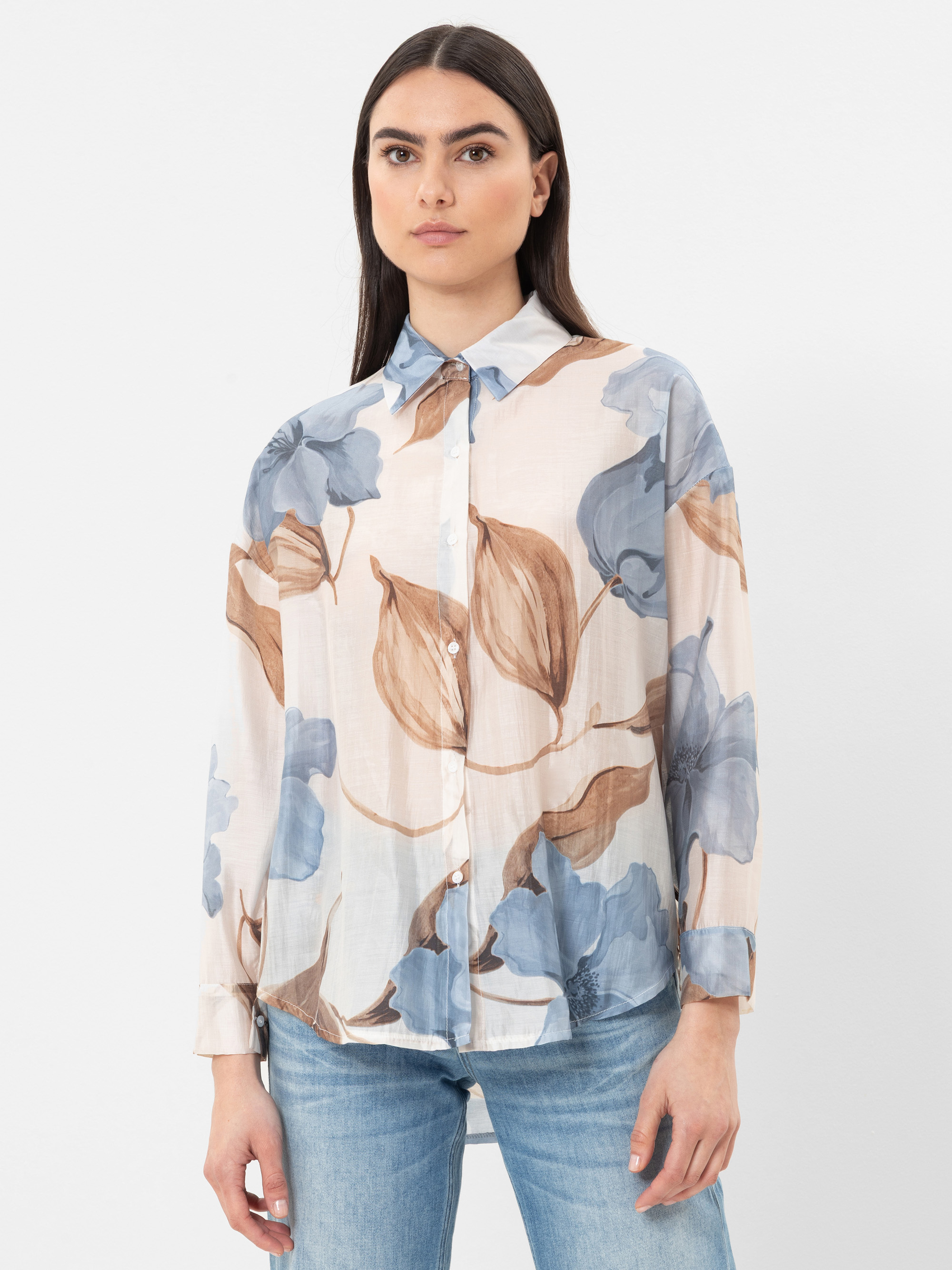 Eine Person, die eine helle, halbdurchsichtige Button-up-Bluse mit einem großen Blumendruck in Blau- und Brauntönen trägt, kombiniert mit einer hellblauen Jeans, steht vor einem schlichten weißen Hintergrund.