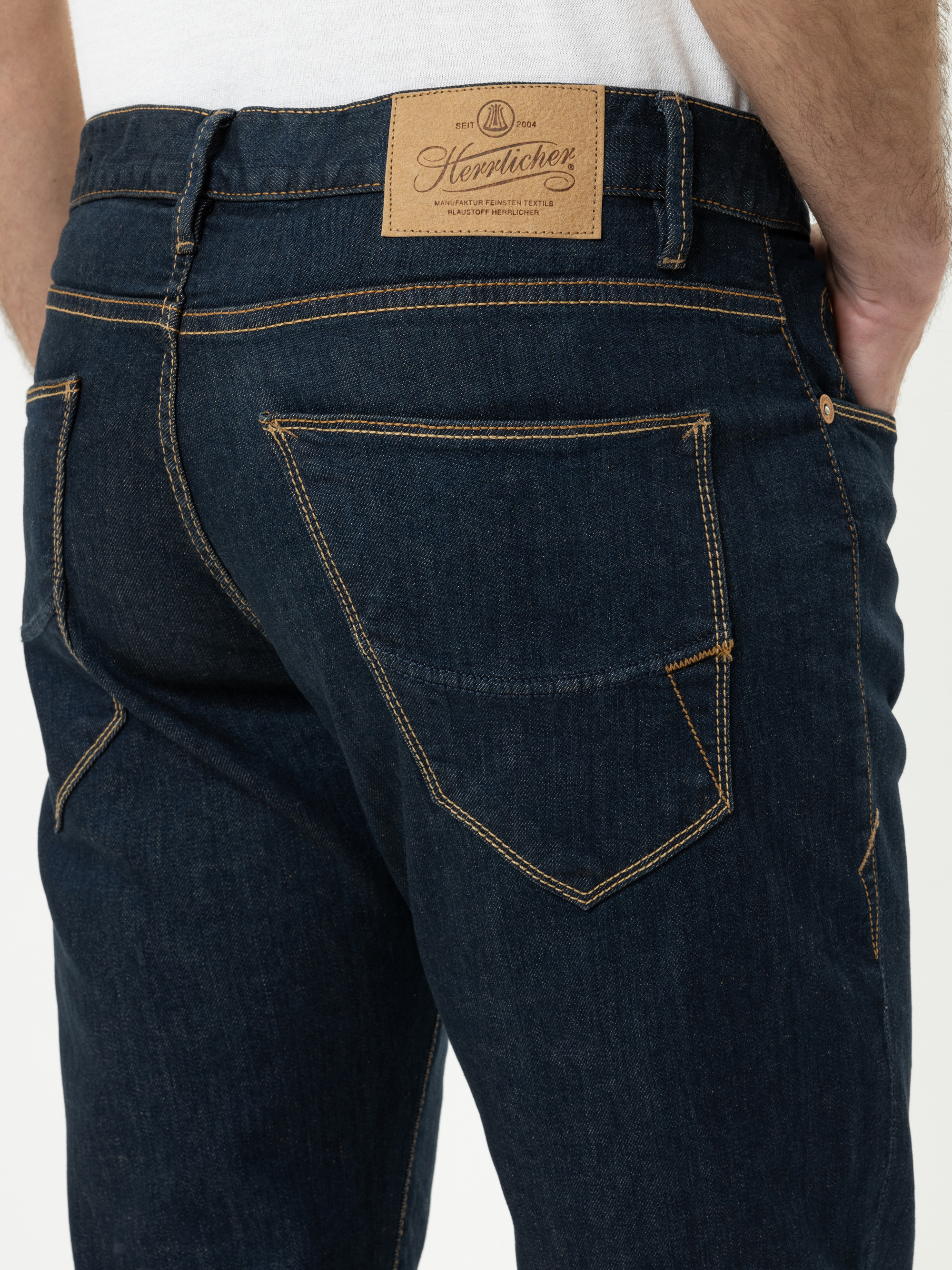 Nahaufnahme einer Person, die eine dunkelblaue Jeans mit Kontrastnähten trägt, wobei die Gesäßtaschen und ein brauner Lederaufnäher mit dem Herrlicher-Markennamen am Bund zu sehen sind.