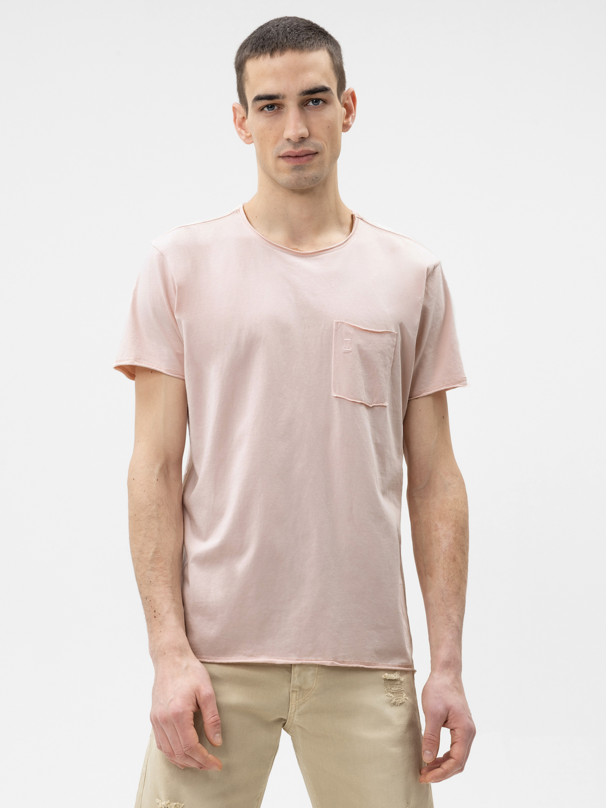 Ein junger Mann mit kurzen dunklen Haaren trägt ein hellrosa T-Shirt mit einer Brusttasche und eine beigefarbene Hose. Er steht mit dem Gesicht nach vorne vor einem schlichten weißen Hintergrund.