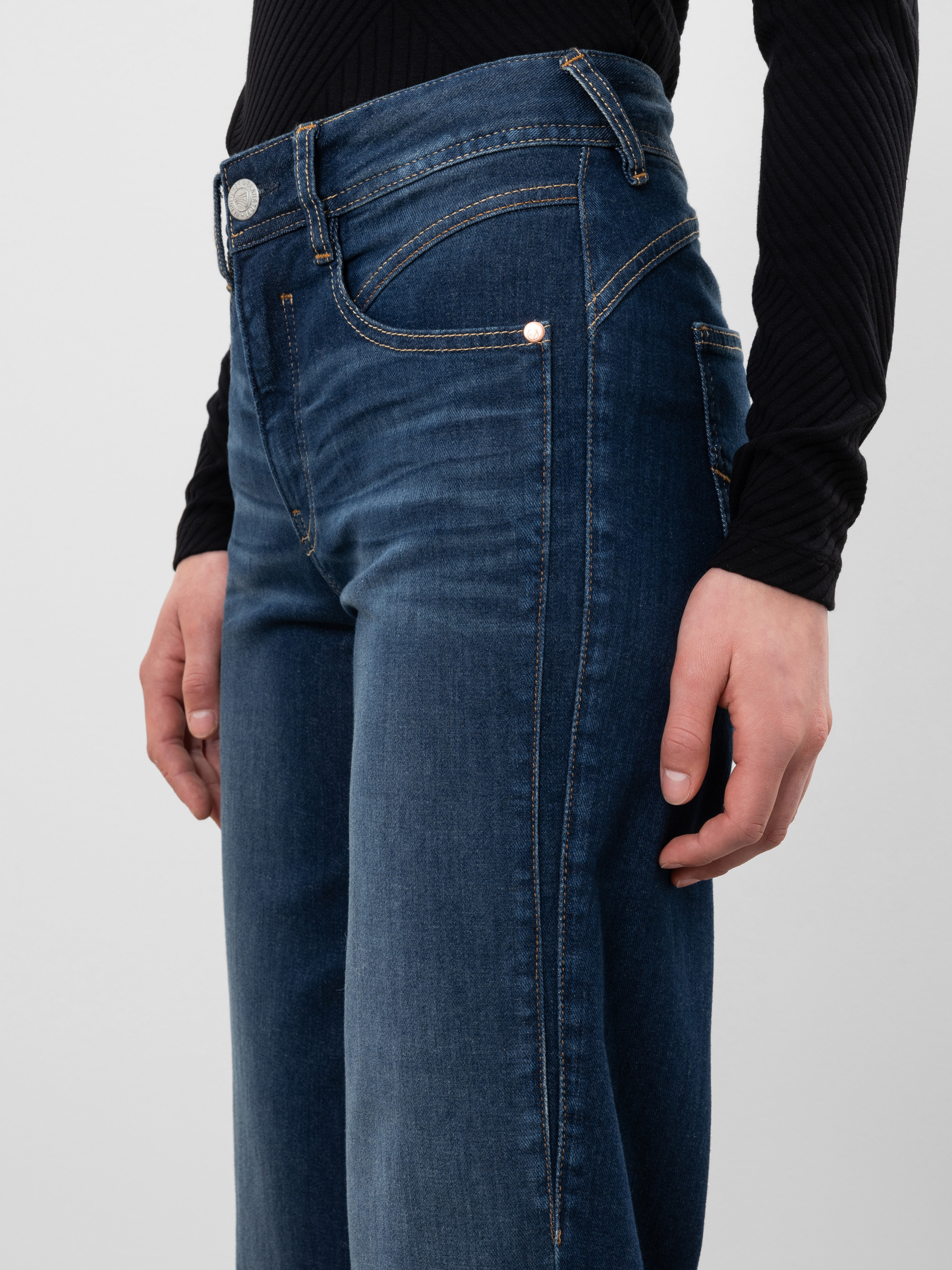 Eine Person trägt eine dunkelblaue Jeans mit hoher Taille und ein schwarzes, langärmeliges Oberteil. Das Bild konzentriert sich auf die Jeans und zeigt die Vordertaschen, den Bund und einen Teil der Hände der Person.