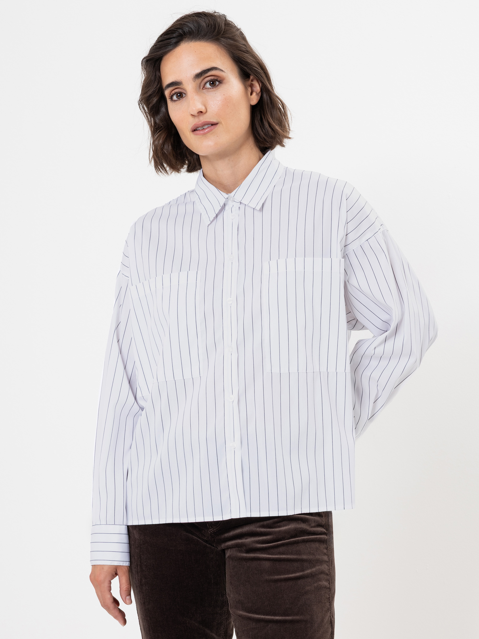 Eine Person mit schulterlangen braunen Haaren trägt ein weißes Button-up-Hemd mit dünnen vertikalen schwarzen Streifen und eine braune Kordhose vor einem schlichten weißen Hintergrund.