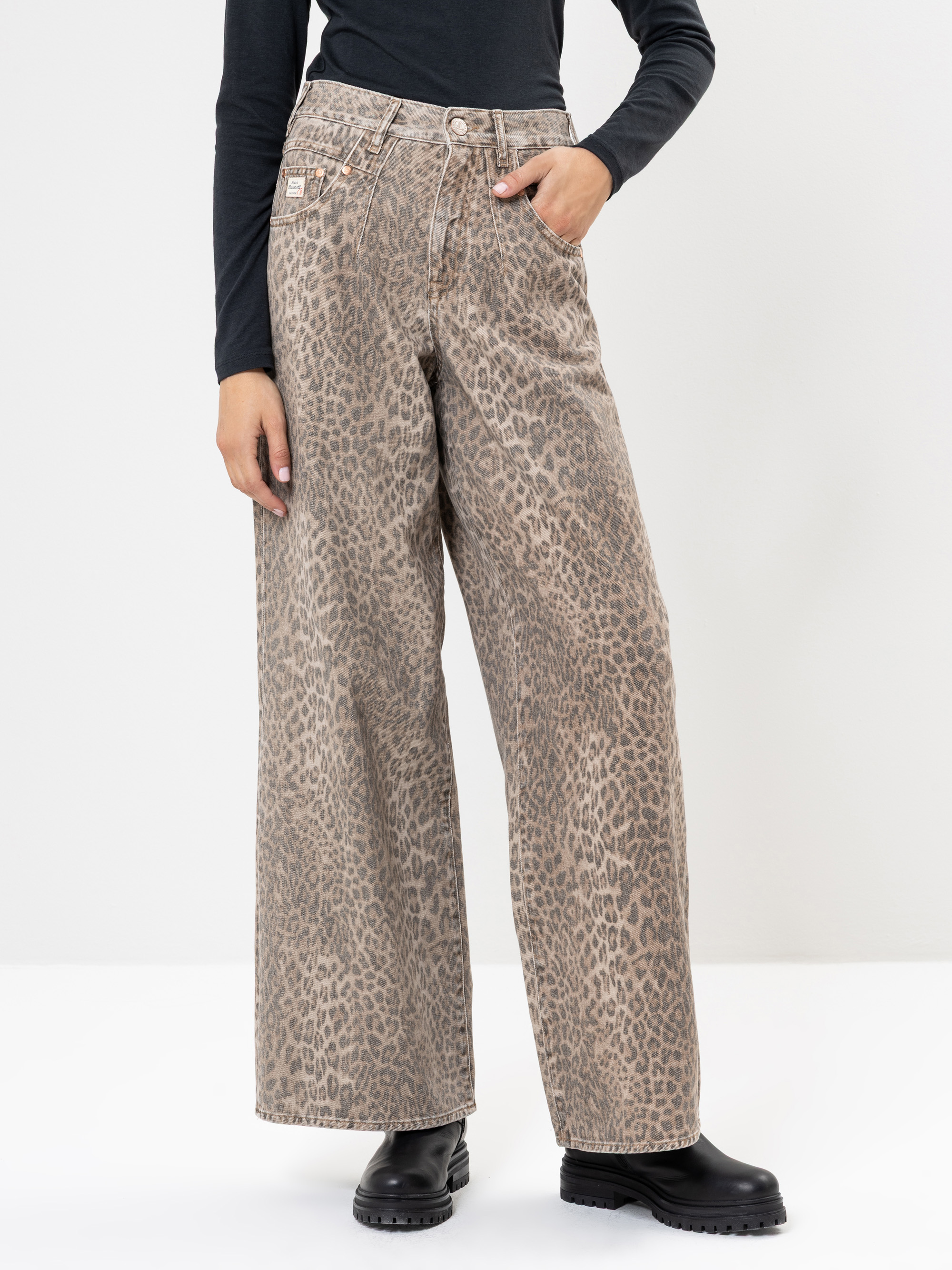 Eine Person, die eine hochtaillierte Hose mit weitem Bein und Leopardenmuster, ein schwarzes, langärmeliges Oberteil und schwarze, klobige Stiefel trägt, steht vor einem schlichten weißen Hintergrund und hat eine Hand in einer Tasche.