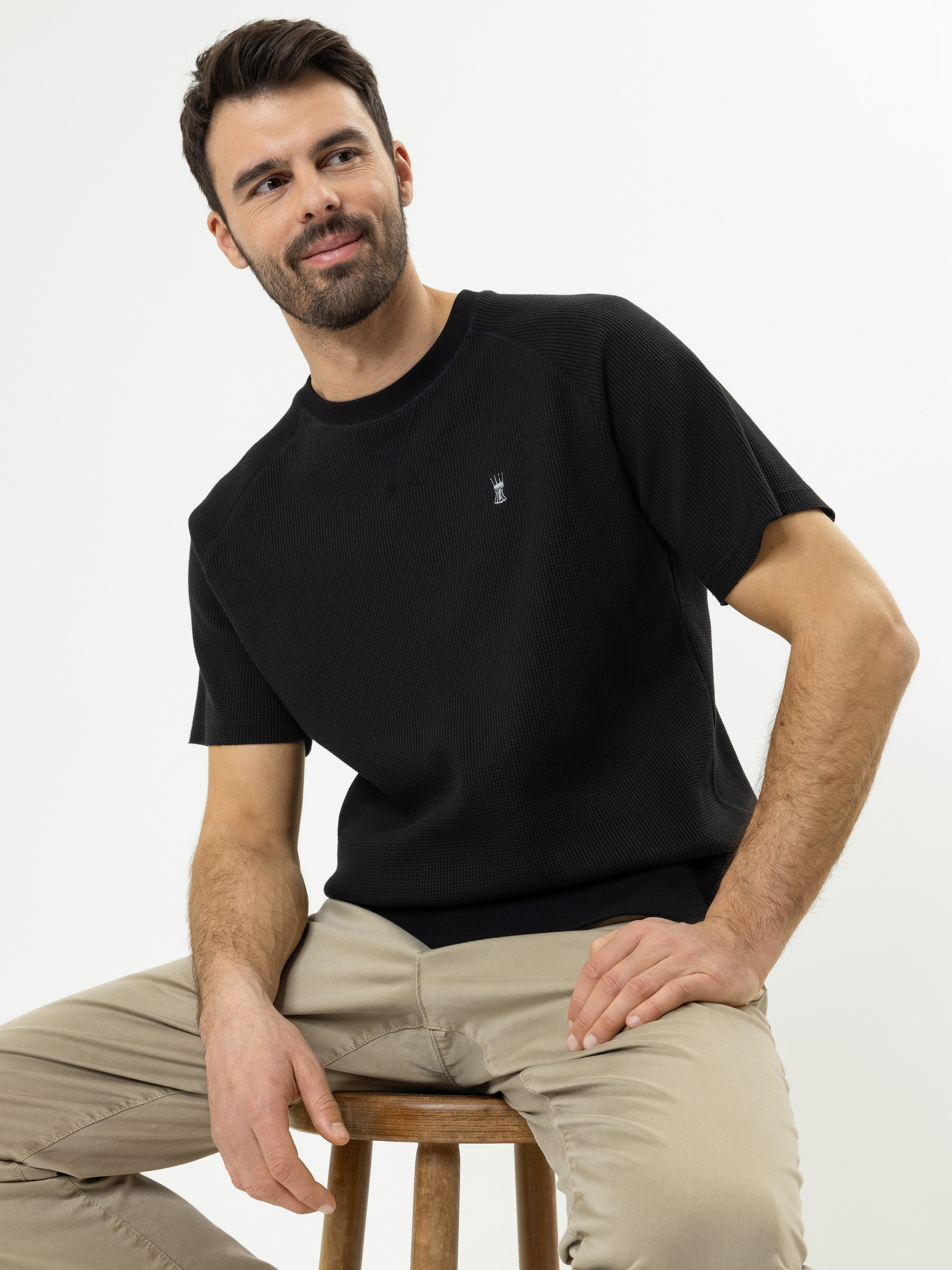 Ein Mann mit kurzen dunklen Haaren und Bart sitzt auf einem Holzschemel, trägt ein schwarzes kurzärmeliges Hemd und eine beigefarbene Hose, lächelt leicht und blickt zur Seite vor einem schlichten weißen Hintergrund.