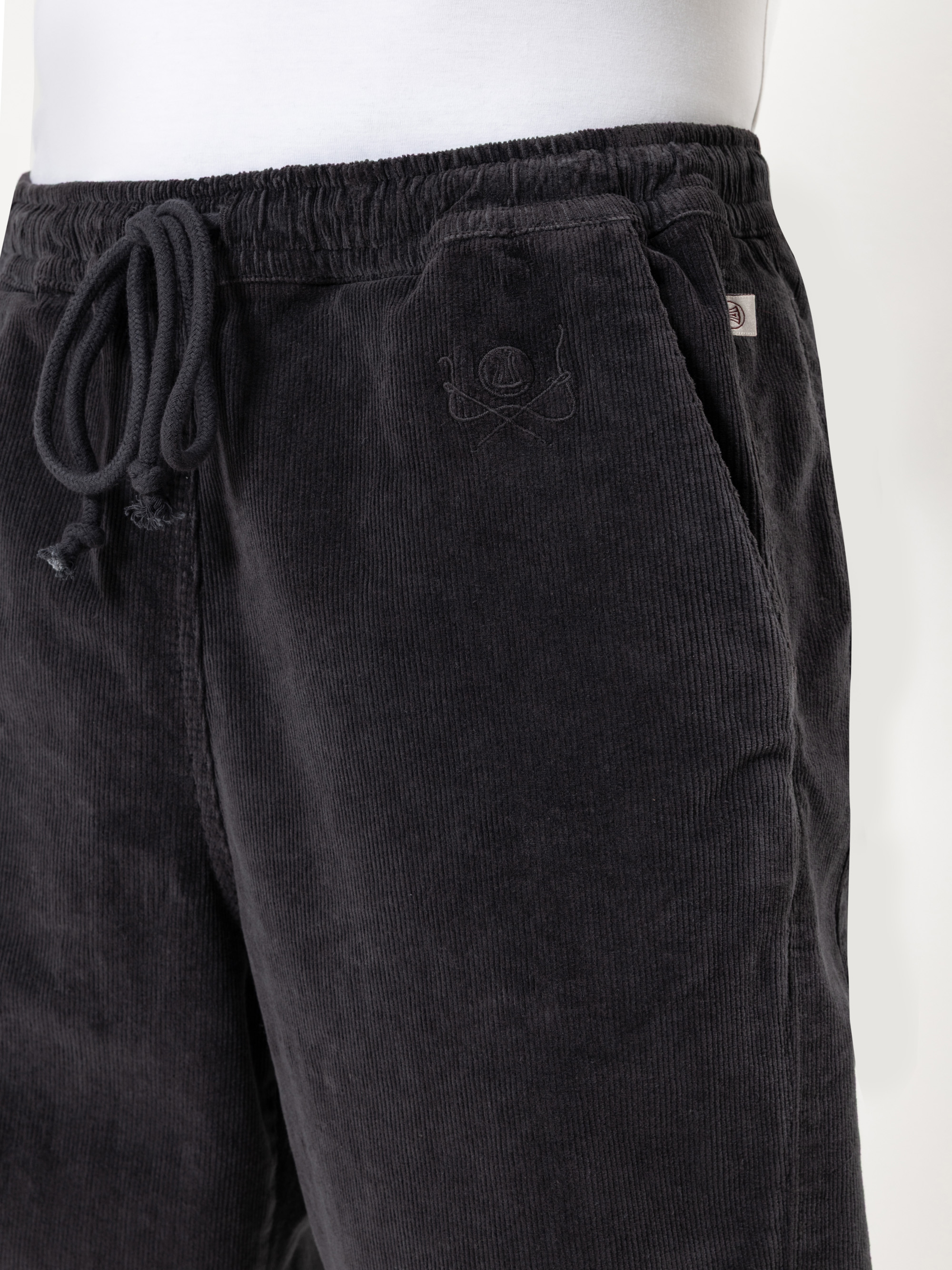 Nahaufnahme einer Person, die eine schwarze Kordhose mit elastischem Bund und Kordelzug trägt; eine Hand steckt in der Tasche. Die Hose ist in der Nähe der Tasche mit einer dezenten Stickerei versehen.