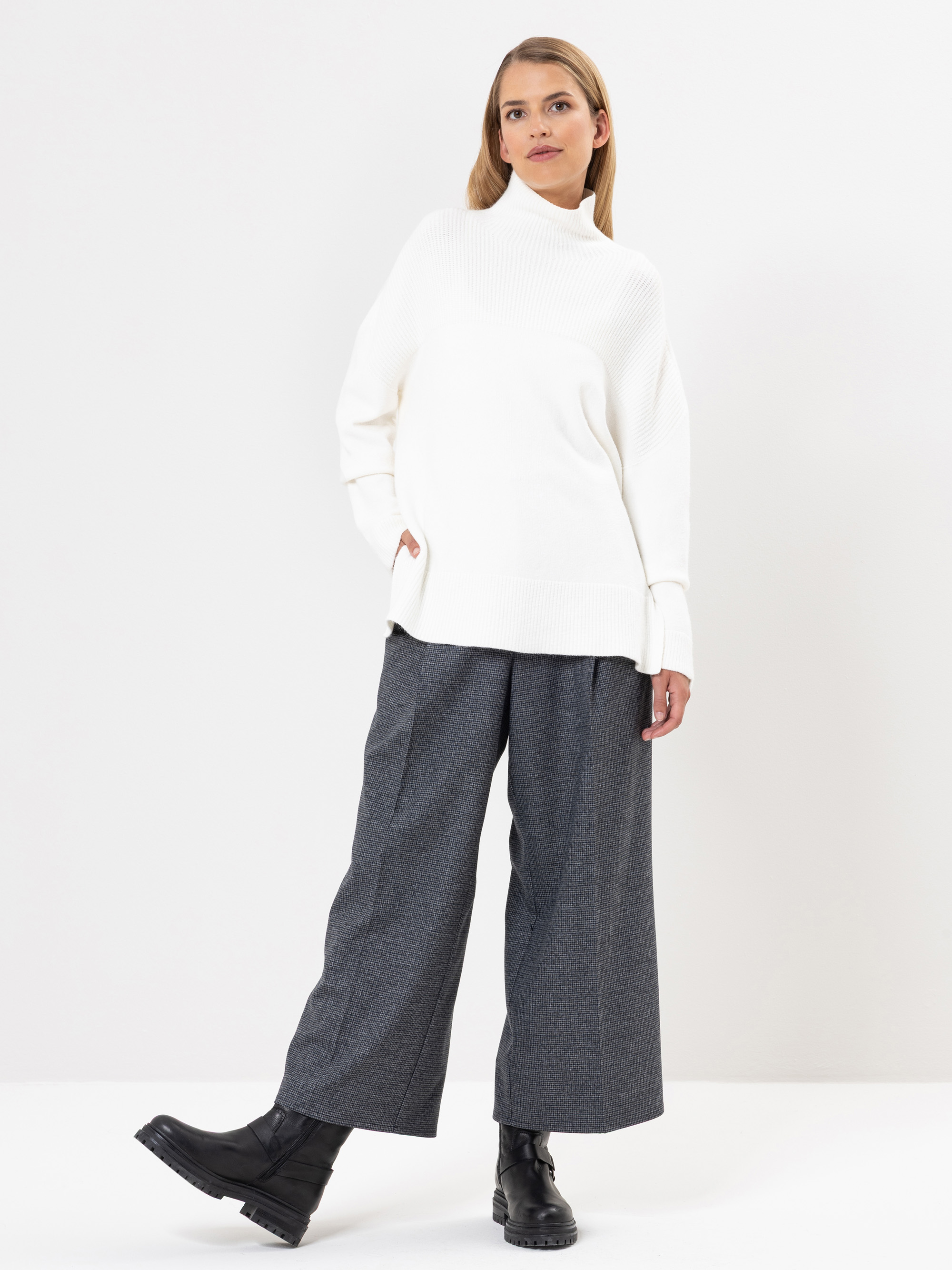 Eine Frau posiert vor einem weißen Hintergrund und trägt einen weißen Rollkragenpullover, eine graue Hose mit weitem Bein und schwarze Stiefel. Sie steht mit einer Hand in der Tasche und einem Fuß leicht nach vorne.