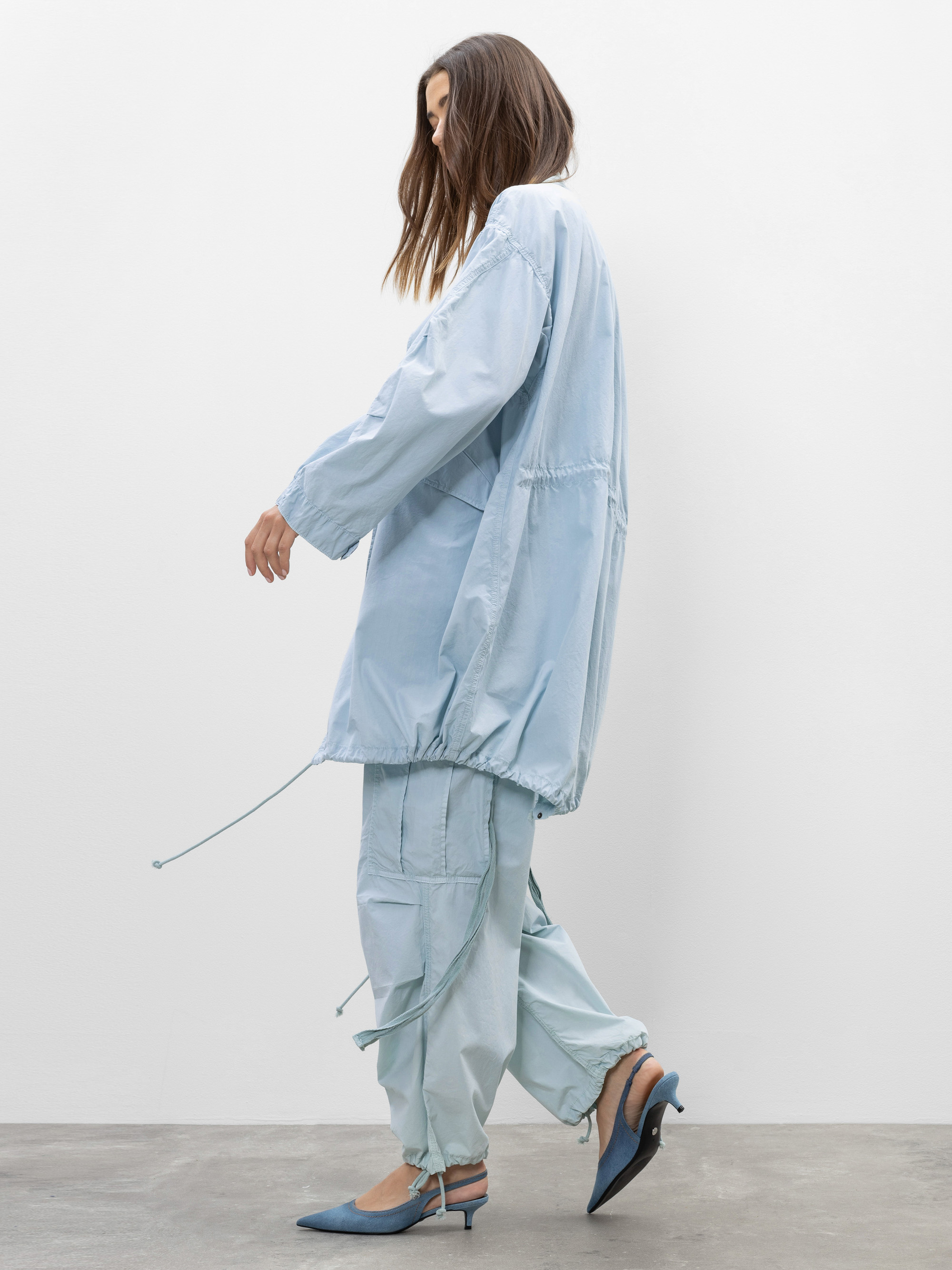 Eine Frau, die eine lockere hellblaue Jacke und eine dazu passende Schlabberhose mit Kordelzug trägt, kombiniert mit blauen spitzen Absätzen, geht im Profil vor einem schlichten weißen Hintergrund.