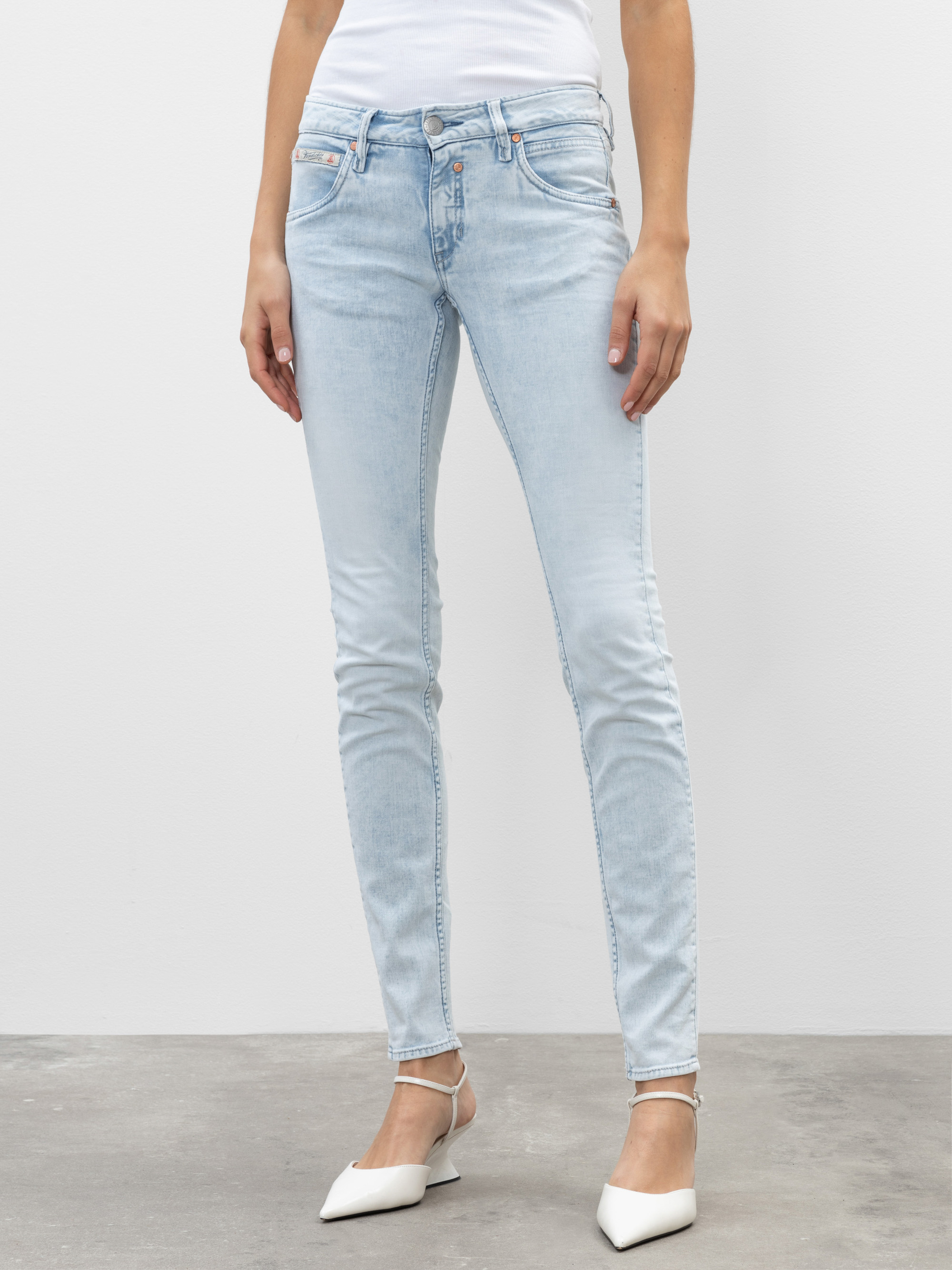 Eine Person, die hellblaue Skinny Jeans und weiße spitze Absätze trägt, steht vor einem schlichten weißen Hintergrund. Die Jeans hat ein klassisches Five-Pocket-Design und eine leicht verblasste Waschung.