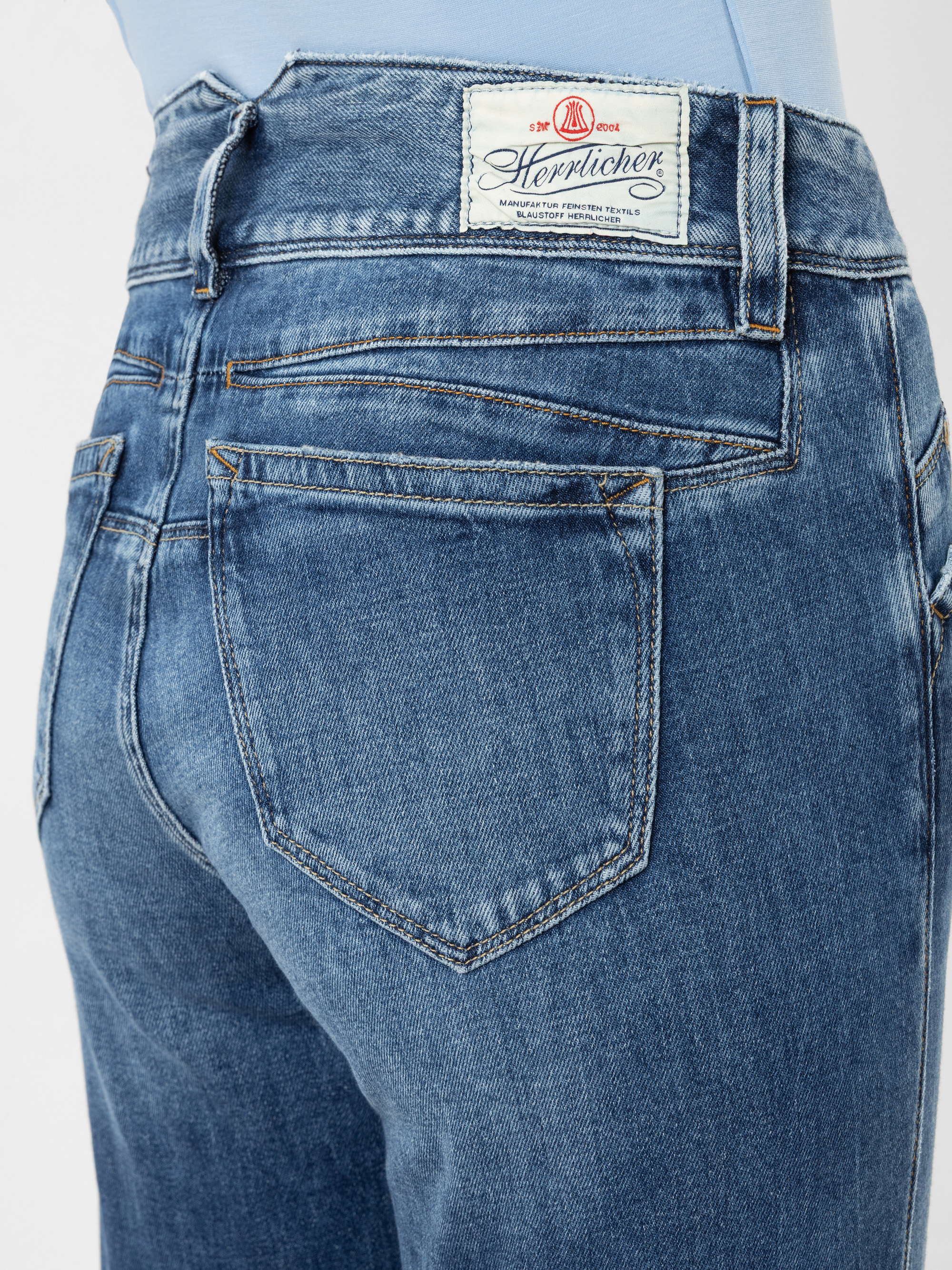 Nahaufnahme der Rückenansicht einer blauen Denim-Jeans mit zwei Gesäßtaschen, sichtbaren Nähten und einem weißen Herrlicher-Markenetikett über der rechten Tasche. Die Person trägt ein hellblaues Hemd, das in die Hose gesteckt ist.